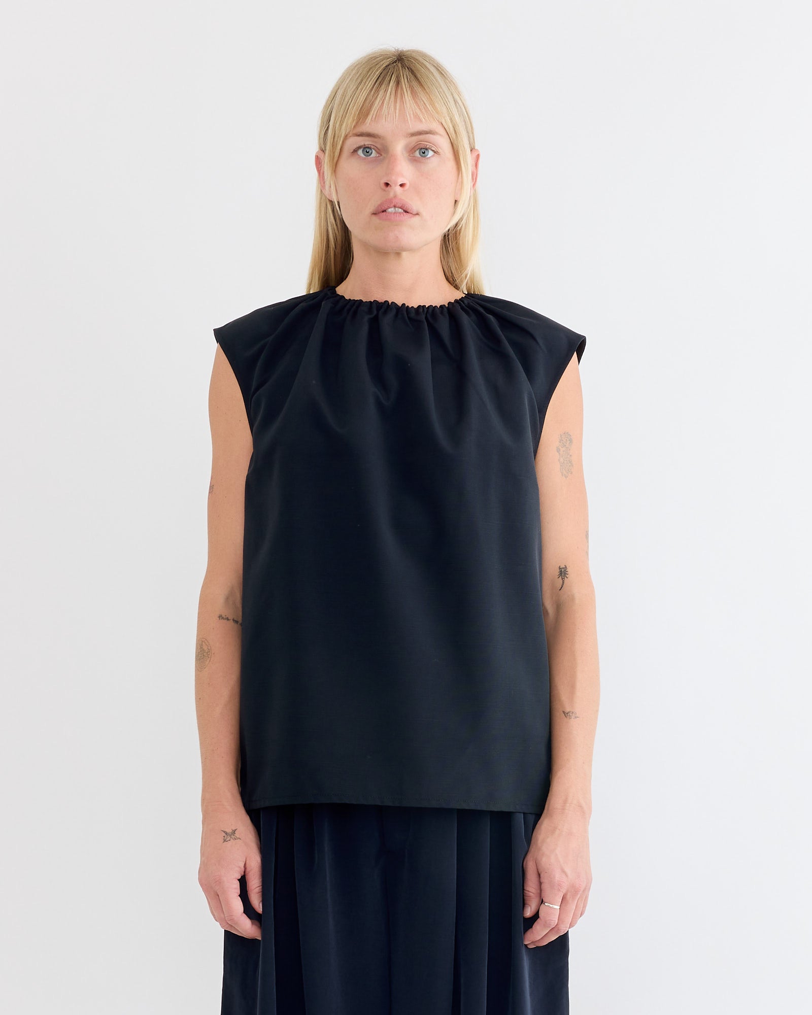 A person with straight blonde hair and bangs stands facing forward, wearing the Tibi Lotte Nylon Shirred Neck Top in Black and a black skirt against a plain white background. Small arm tattoos are visible.