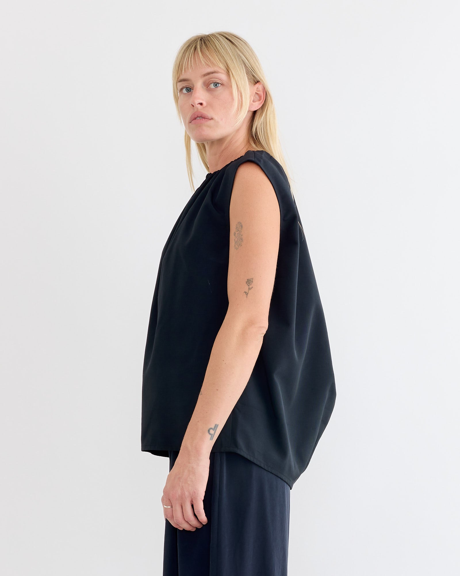 A person with straight blonde hair and visible arm tattoos stands sideways, wearing the Tibi Lotte Nylon Shirred Neck Top in Black and black pants against a plain white background.