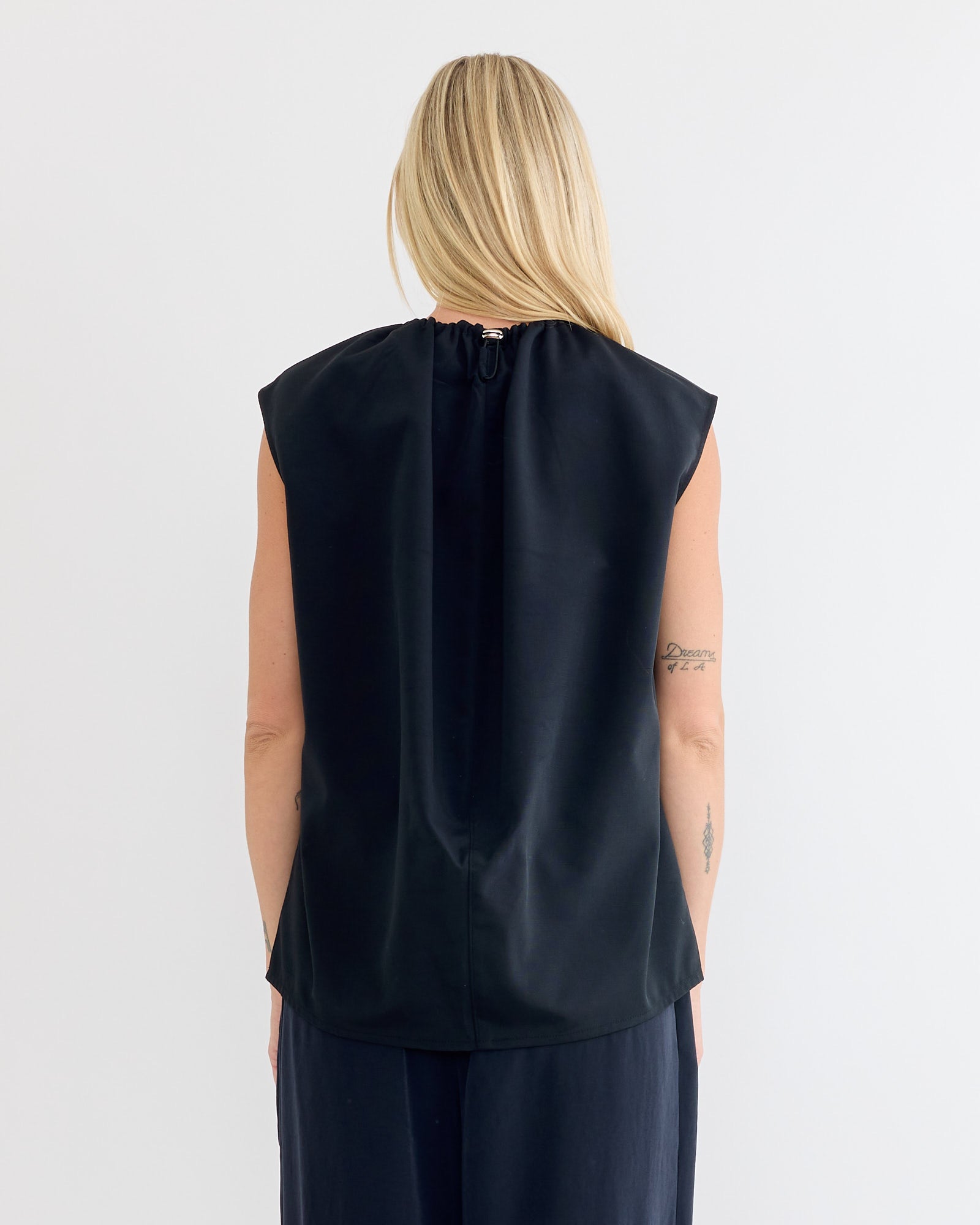 A person with long blonde hair faces away, wearing the Tibi Lotte Nylon Shirred Neck Top in black and dark pants. The sleeveless top features a back zipper. A tattoo is visible on their upper left arm against a plain white background.