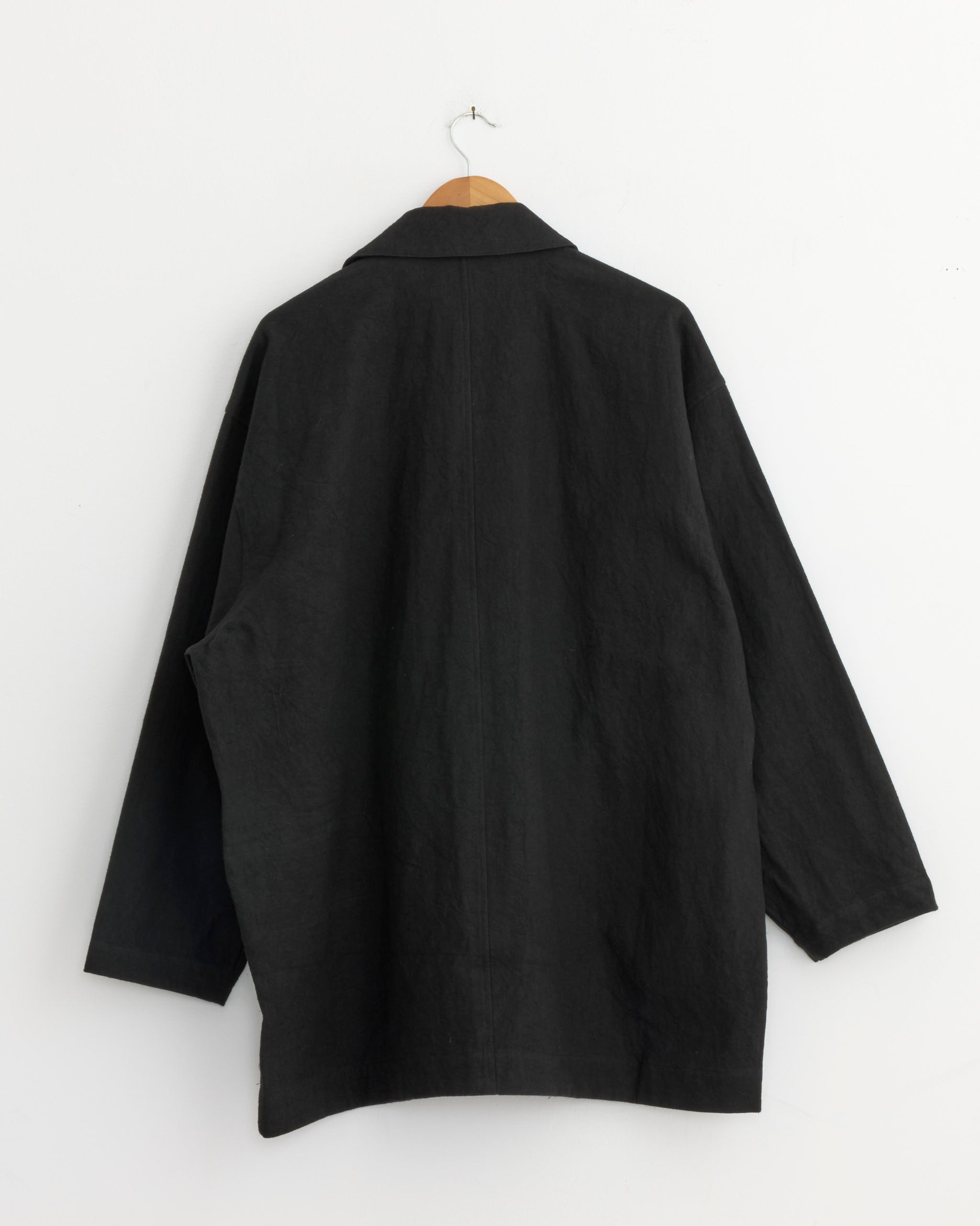 The Cotton Linen Half Coat in Black by Still By Hand hangs on a wooden hanger against a white wall, shown from the back. Made in Japan, this sleek jacket offers timeless appeal.
