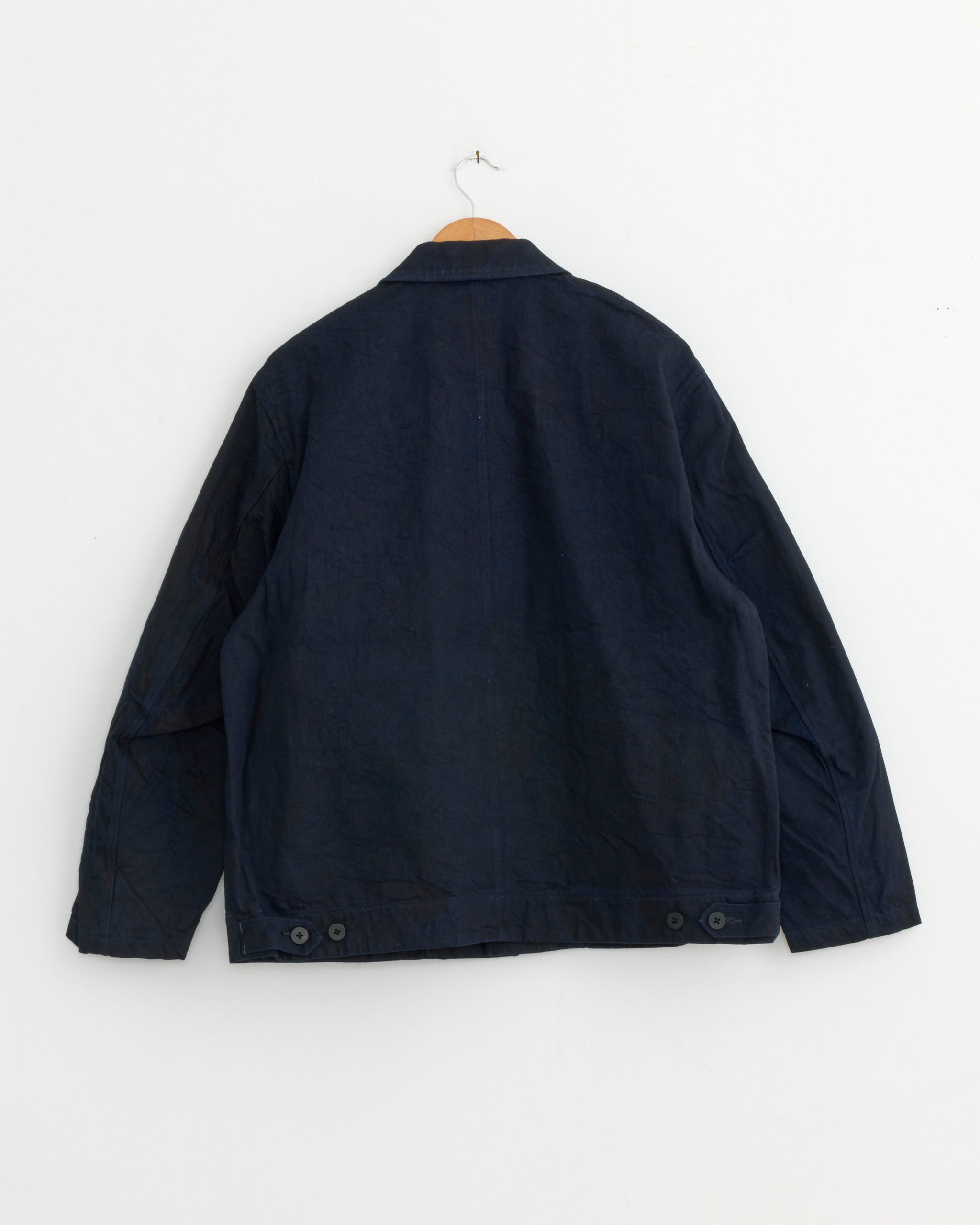 The Still By Hand Denim Jacket in Dark Navy hangs on a wooden hanger against a white wall, displayed from the back and featuring buttons near the bottom hem. Made in Japan.