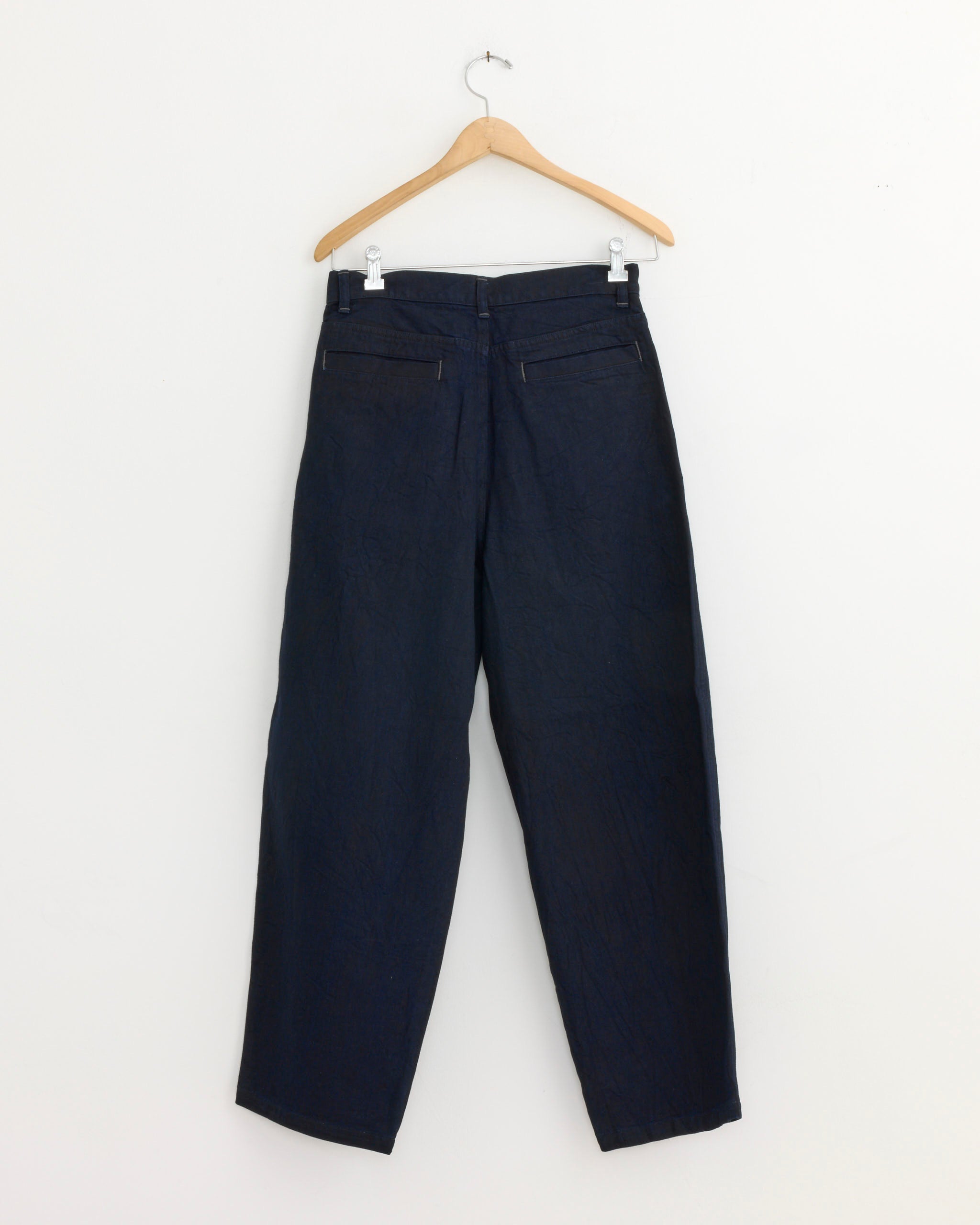 The Still By Hand Deep Tuck Denim Pant in Dark Navy, made in Japan, hangs on a wooden hanger against a plain white wall, displayed from the back to show its pockets and seams.