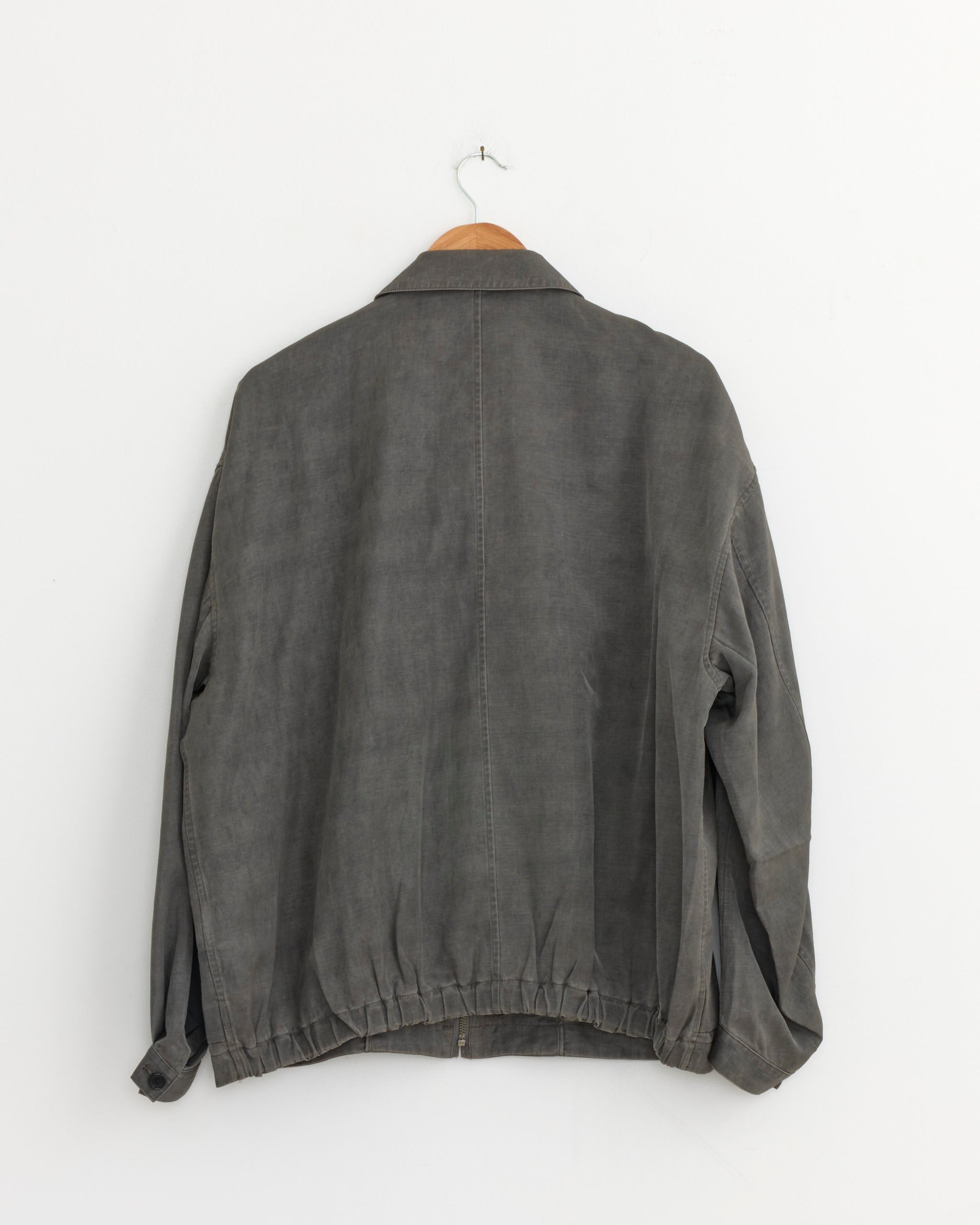 A Pigment Print Zip Up Blouson in Dusty Grey by Still By Hand hangs on a wooden hanger against a white wall, displaying its long sleeves and collar from the back.