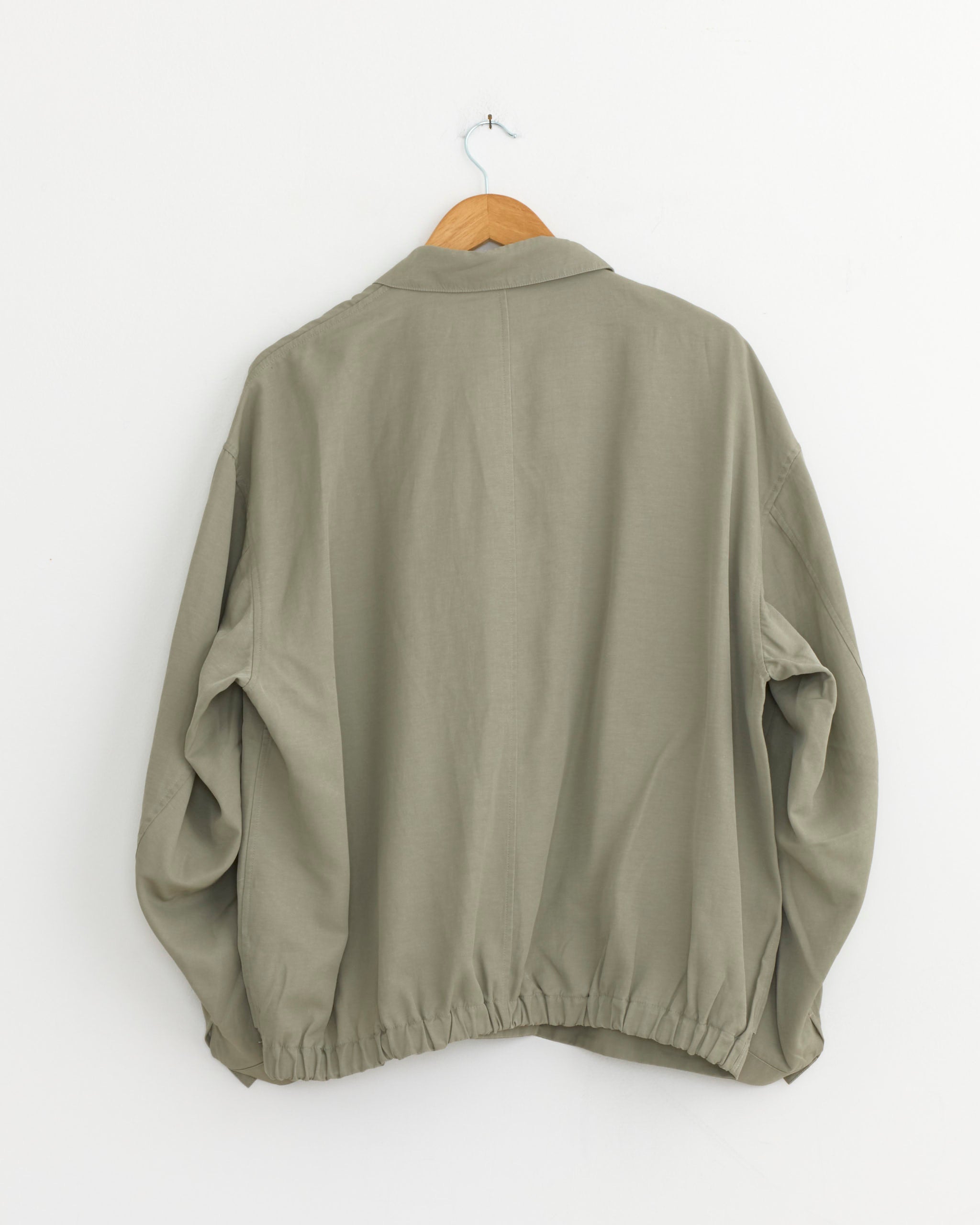 A Still By Hand Pigment Print Zip Up Blouson in Sage hangs on a wooden hanger against a white wall, seen from the back. This loose-fit, long-sleeve jacket with an elasticized hem is made in Japan.