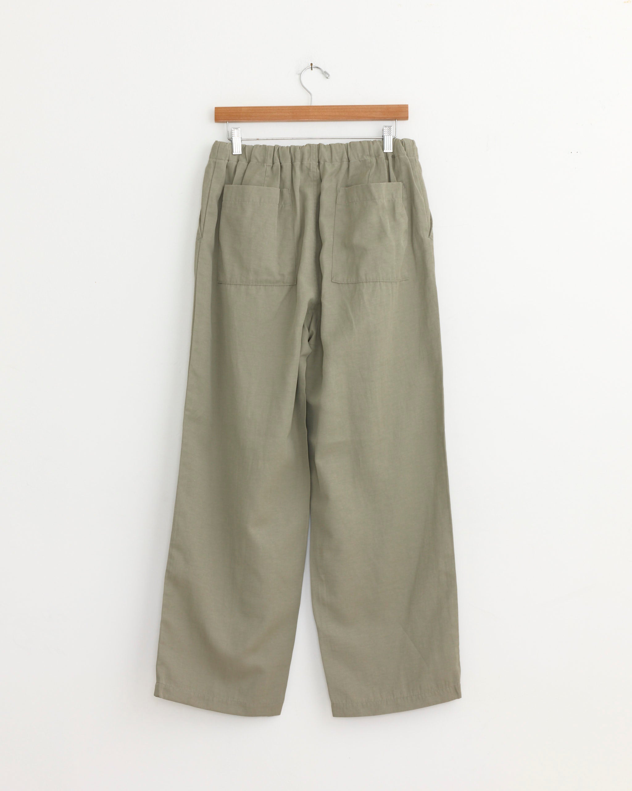 A pair of sage Pigment Print Easy Pants by Still By Hand, made in Japan, with an elastic waistband and two large back pockets, hangs on a wooden hanger against a white wall.