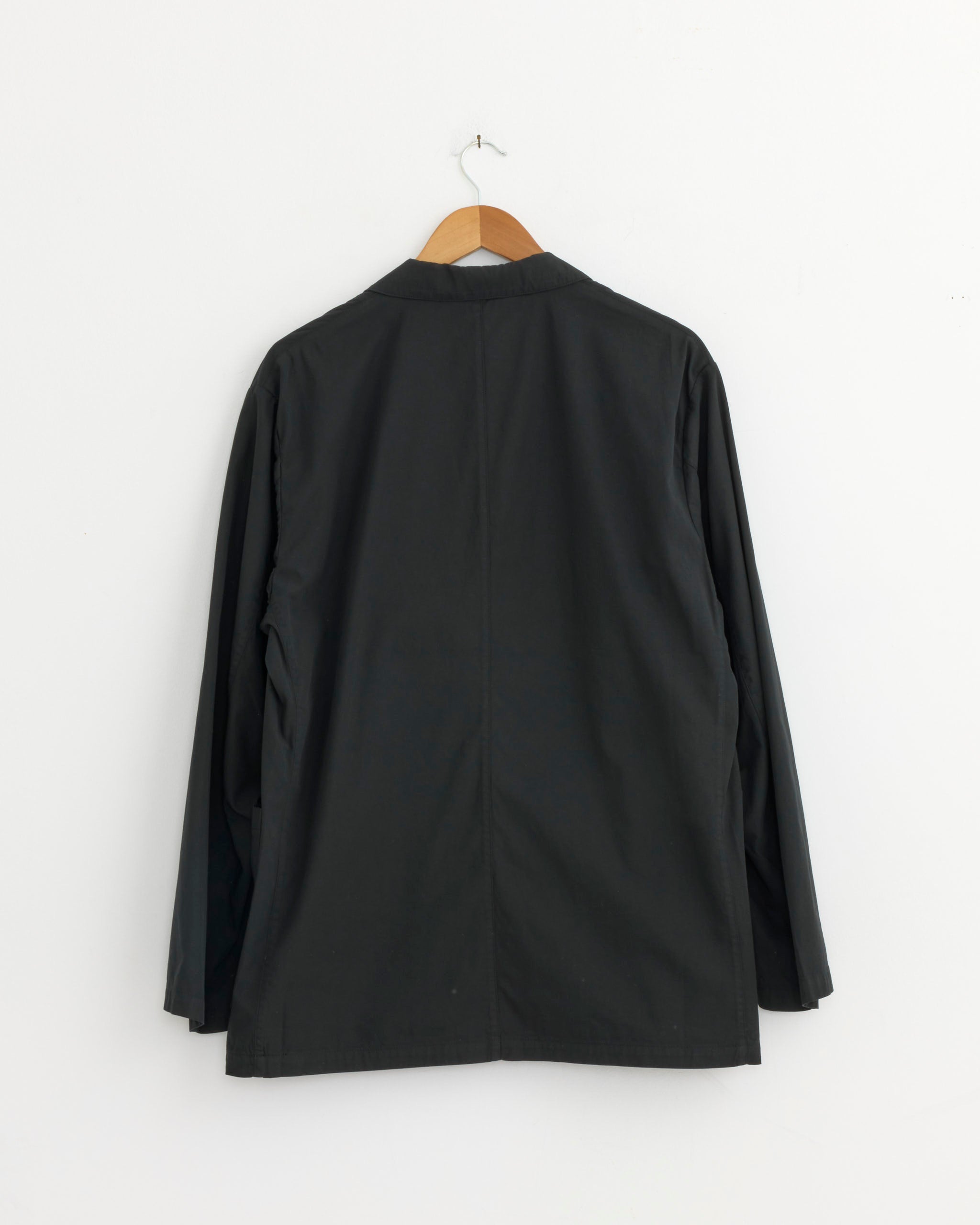 The Still By Hand Garment Dye 3B Jacket in Ink Black, made from high-twist cotton twill in Japan, is shown from the back on a wooden hanger against a plain white wall.