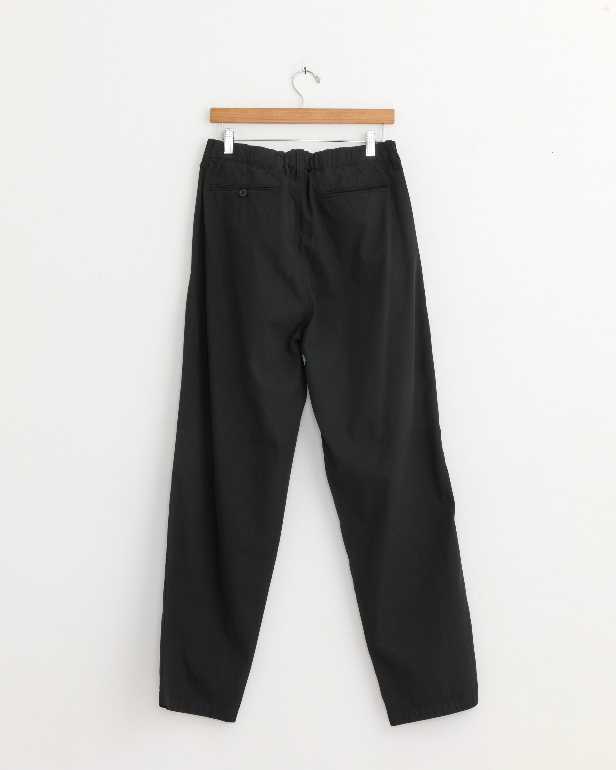 The Still By Hand Garment Dye 2 Pleat Pant in Ink Black, featuring an elastic waistband and back pockets, hangs on a wooden hanger against a plain white wall.