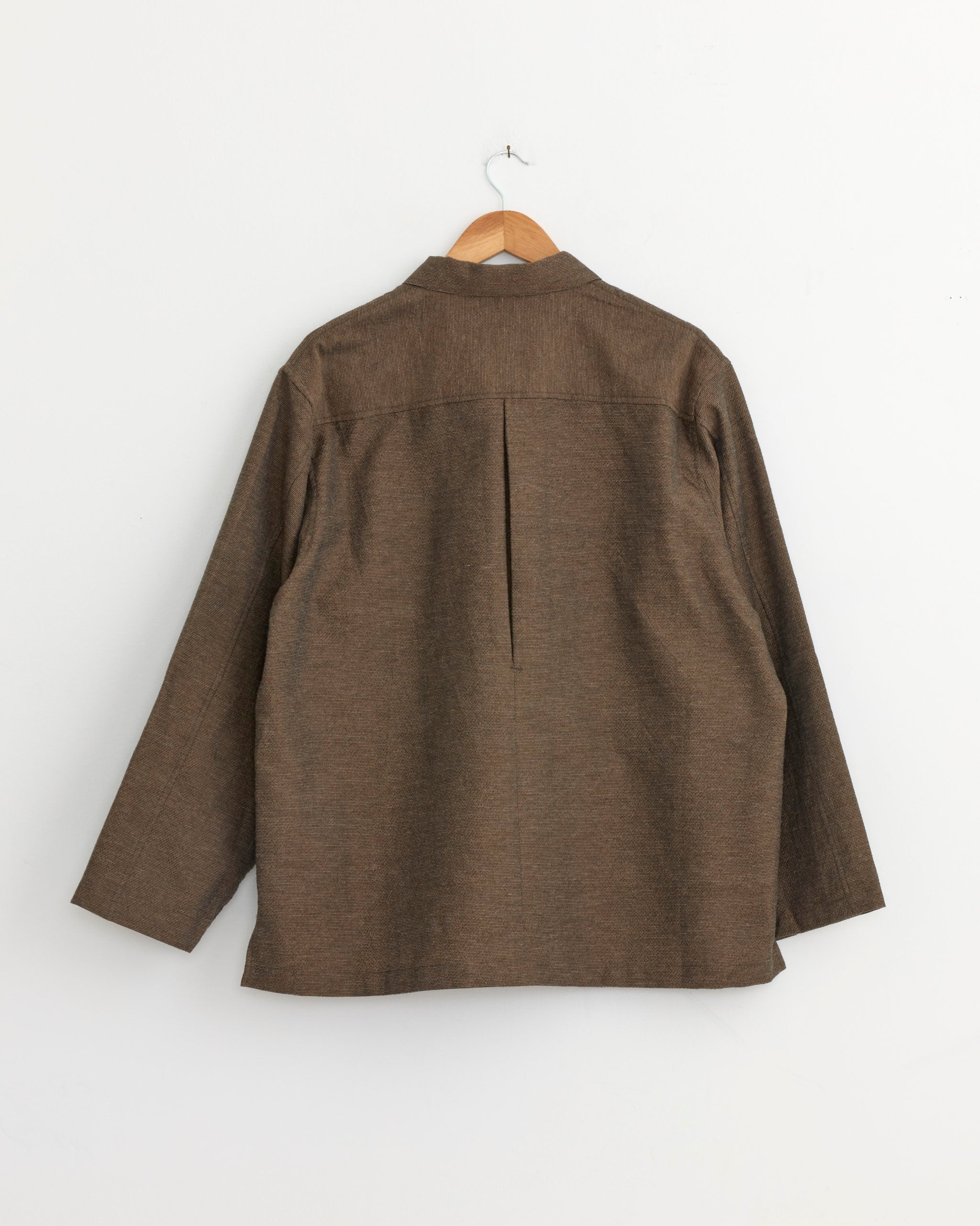The Still By Hand Mixed Color Shirt Jacket in Brown, made from lightweight summer tweed, hangs on a wooden hanger against a white wall and is shown from the back with a single center pleat detail.