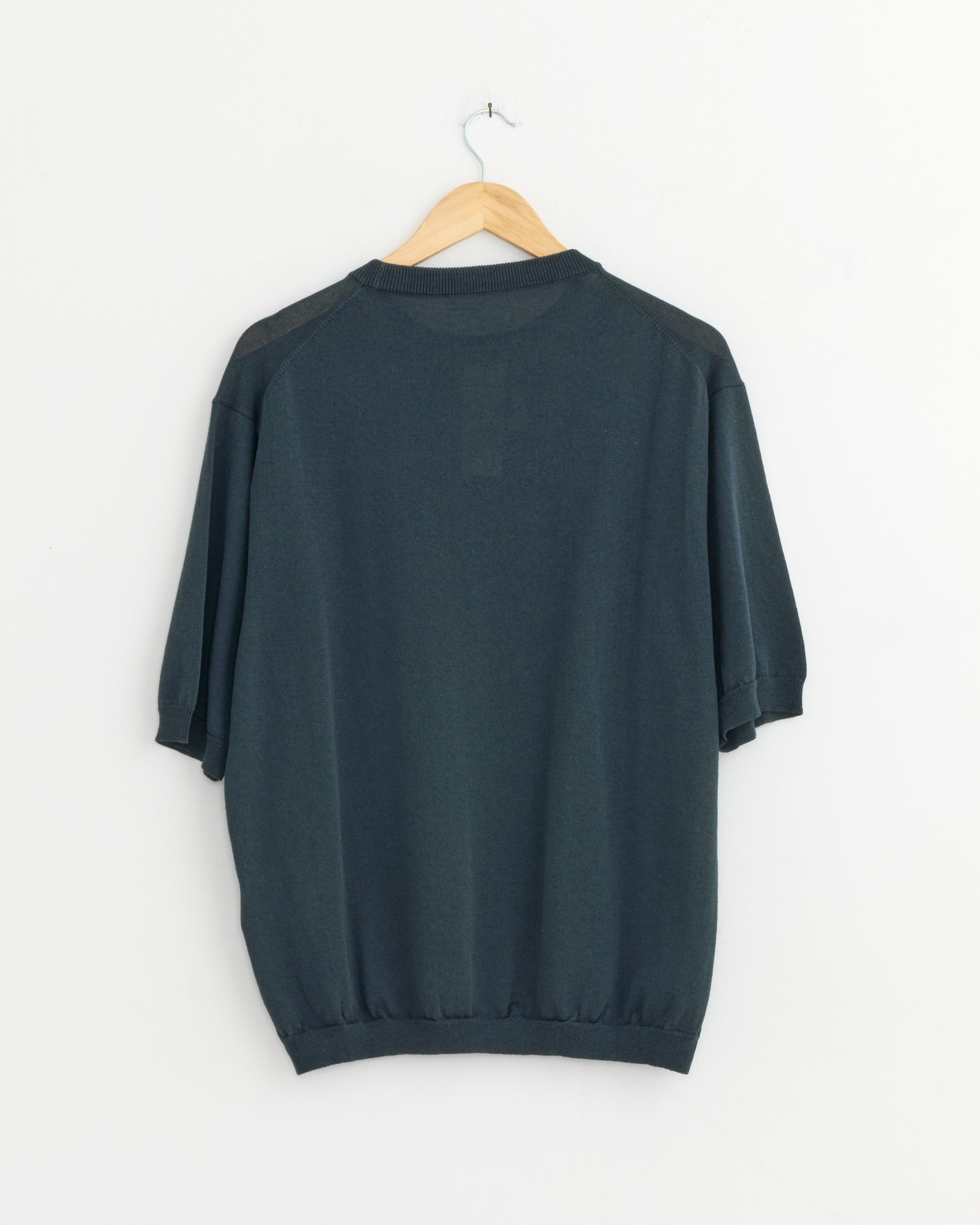 The Half Sleeve Knit T-Shirt in Teal Blue by Still By Hand hangs on a wooden hanger against a plain white wall, displayed from the back.