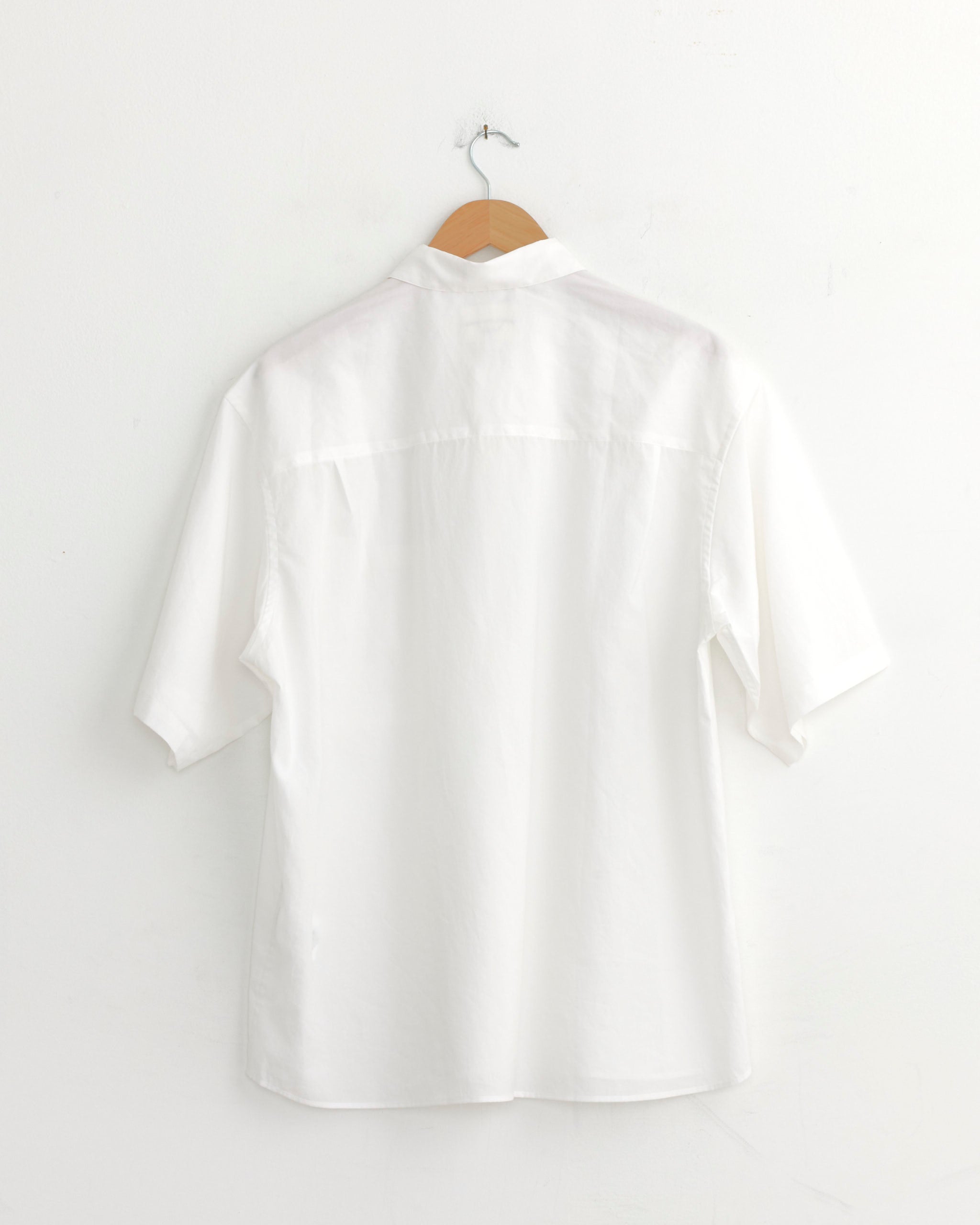 The Still By Hand Half Sleeve Cotton Shirt in White hangs on a wooden hanger against a white wall, showcasing its crisp broadcloth texture from the back view. Made in Japan.