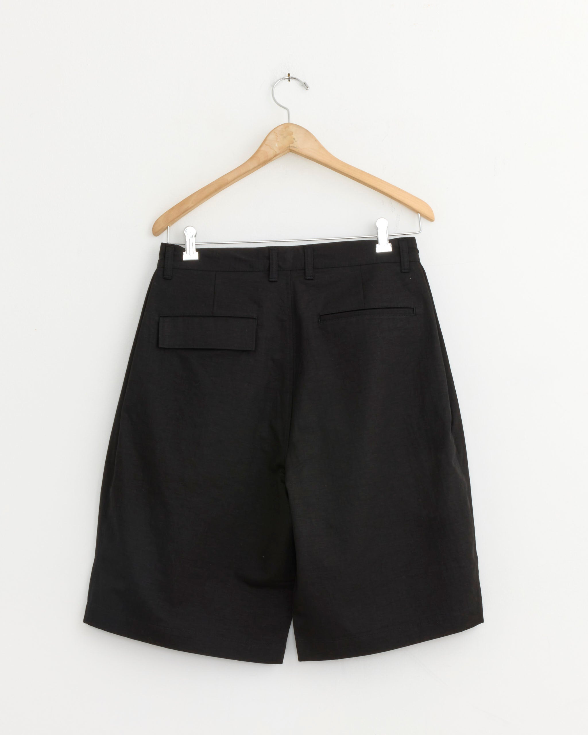 The Still By Hand Linen Mixed Wide Short in Black, crafted in Japan, hangs on a wooden hanger against a plain white wall and is shown from the back, featuring two closed pockets.