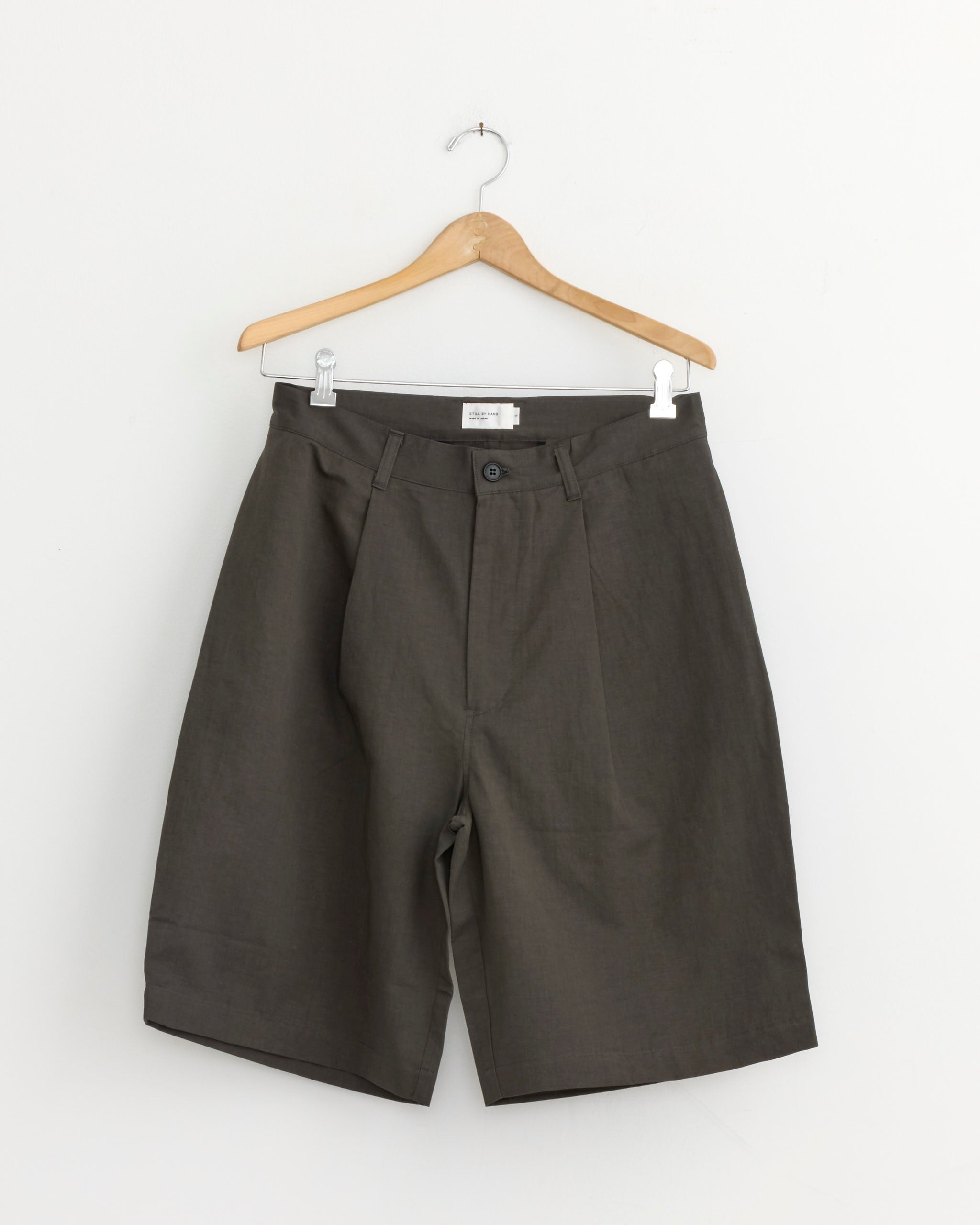 The Linen Mixed Wide Short in Charcoal by Still By Hand hangs on a wooden hanger against a white background, showcasing its button closure, belt loops, and front pleats.