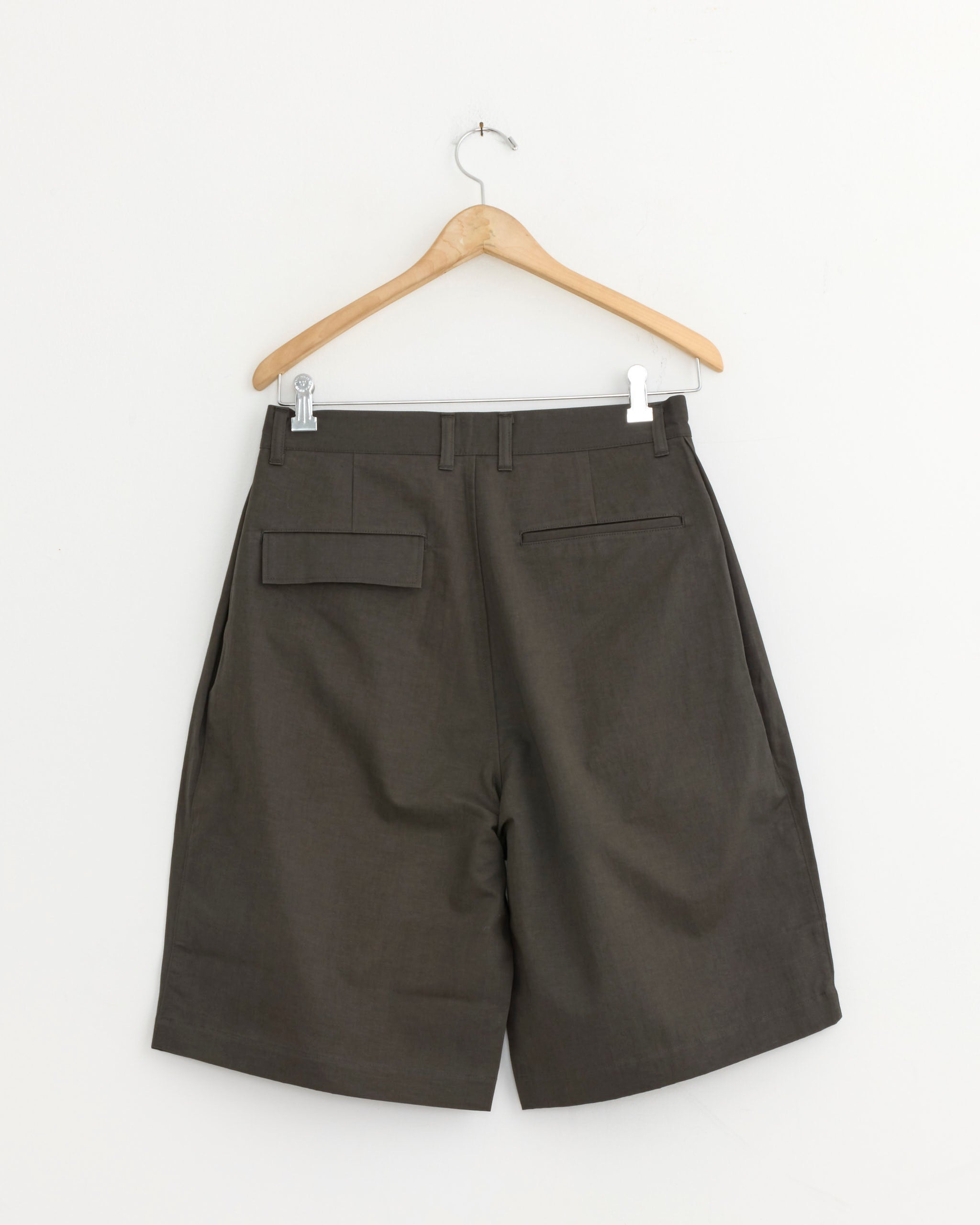 The Still By Hand Linen Mixed Wide Short in Charcoal, made in Japan, hangs on a wooden hanger against a plain white wall, displaying the back view with two pockets.