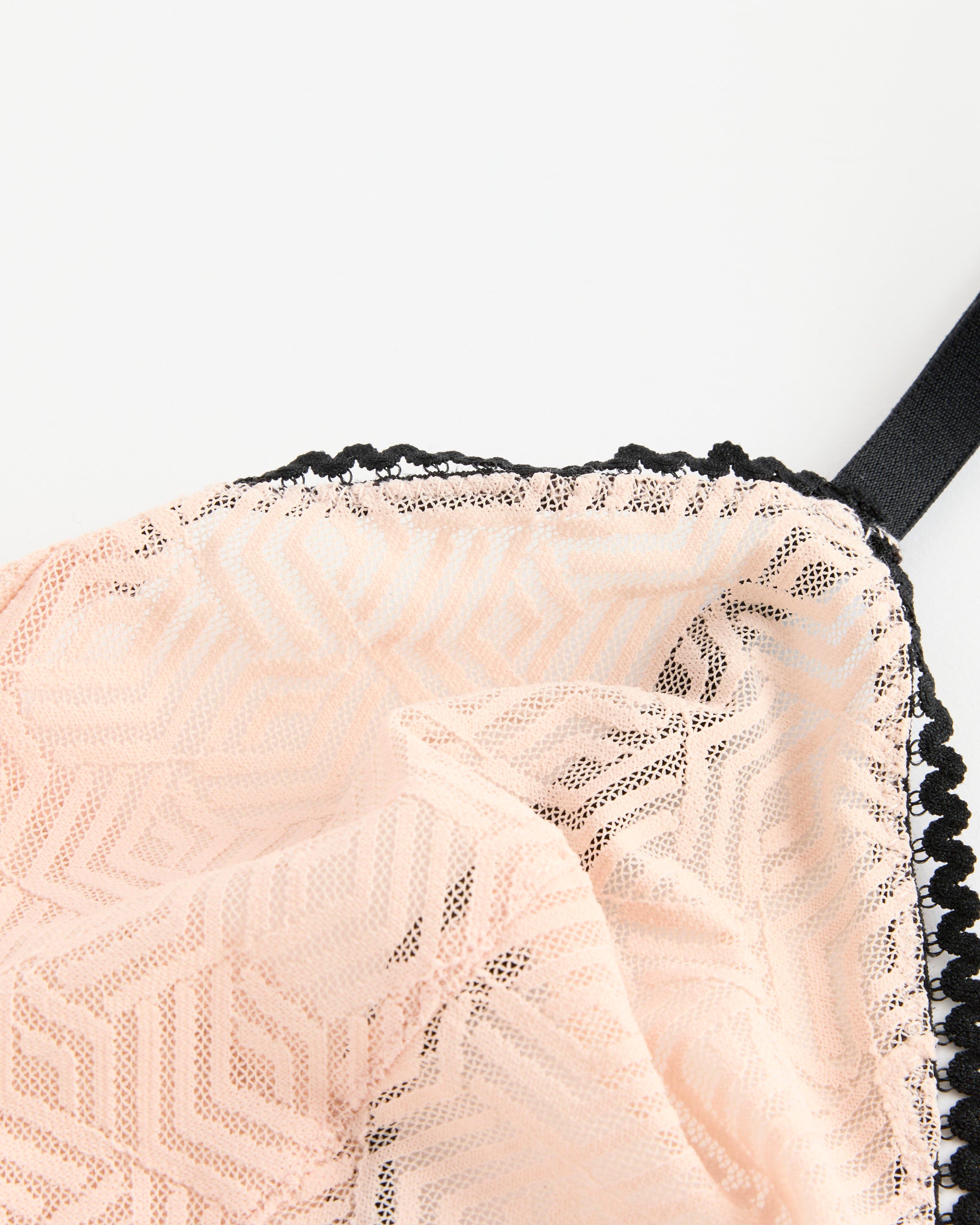 Close-up of the ELSE Synergy Soft Triangle Bra in Soft Pink, featuring geometric lace with black scalloped trim and a black strap. For sizing or fit questions, our online chat is here to help. Displayed on a white background.