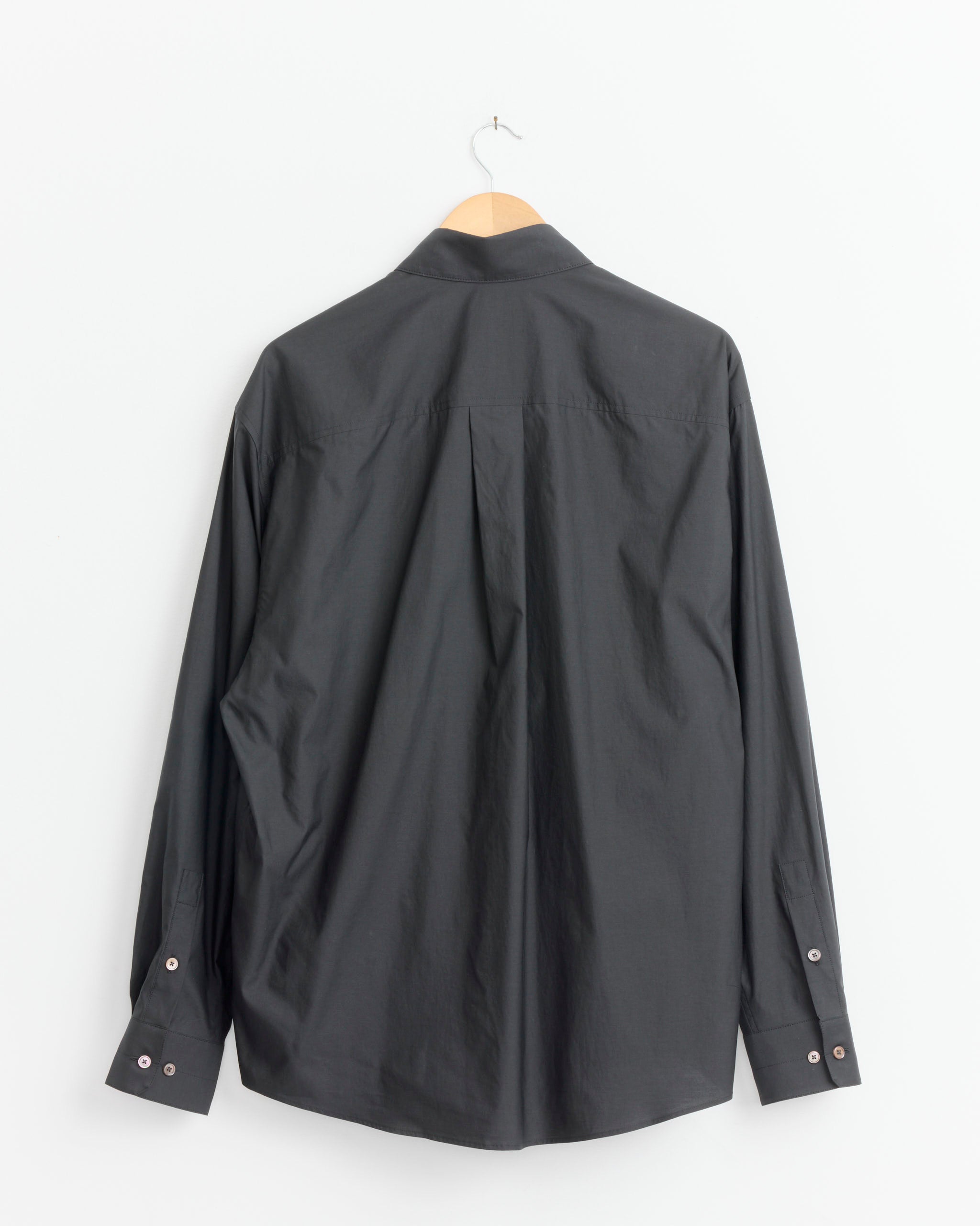 This Lemaire Regular Shirt in Squid Ink, a classic men's long-sleeved style, is displayed on a wooden hanger against a plain white wall, viewed from the back.