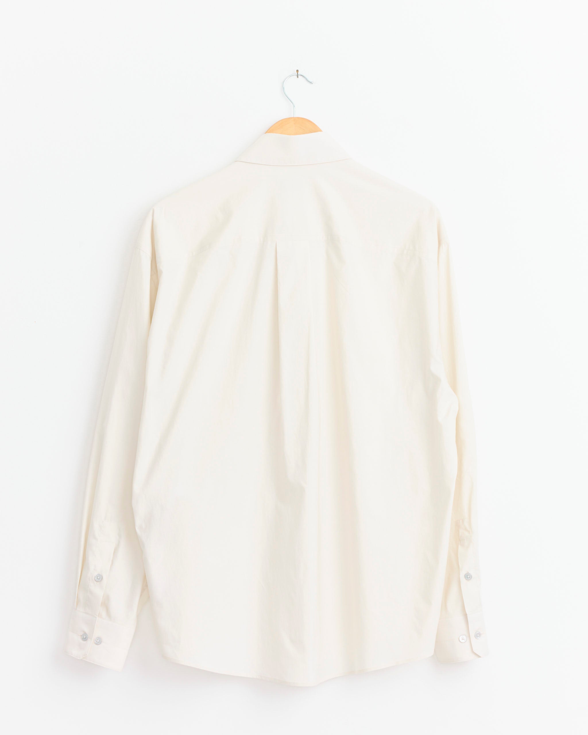 The Lemaire Regular Shirt in Light Cream, crafted from a cotton silk blend, hangs on a wooden hanger against a white background and is shown from the back.