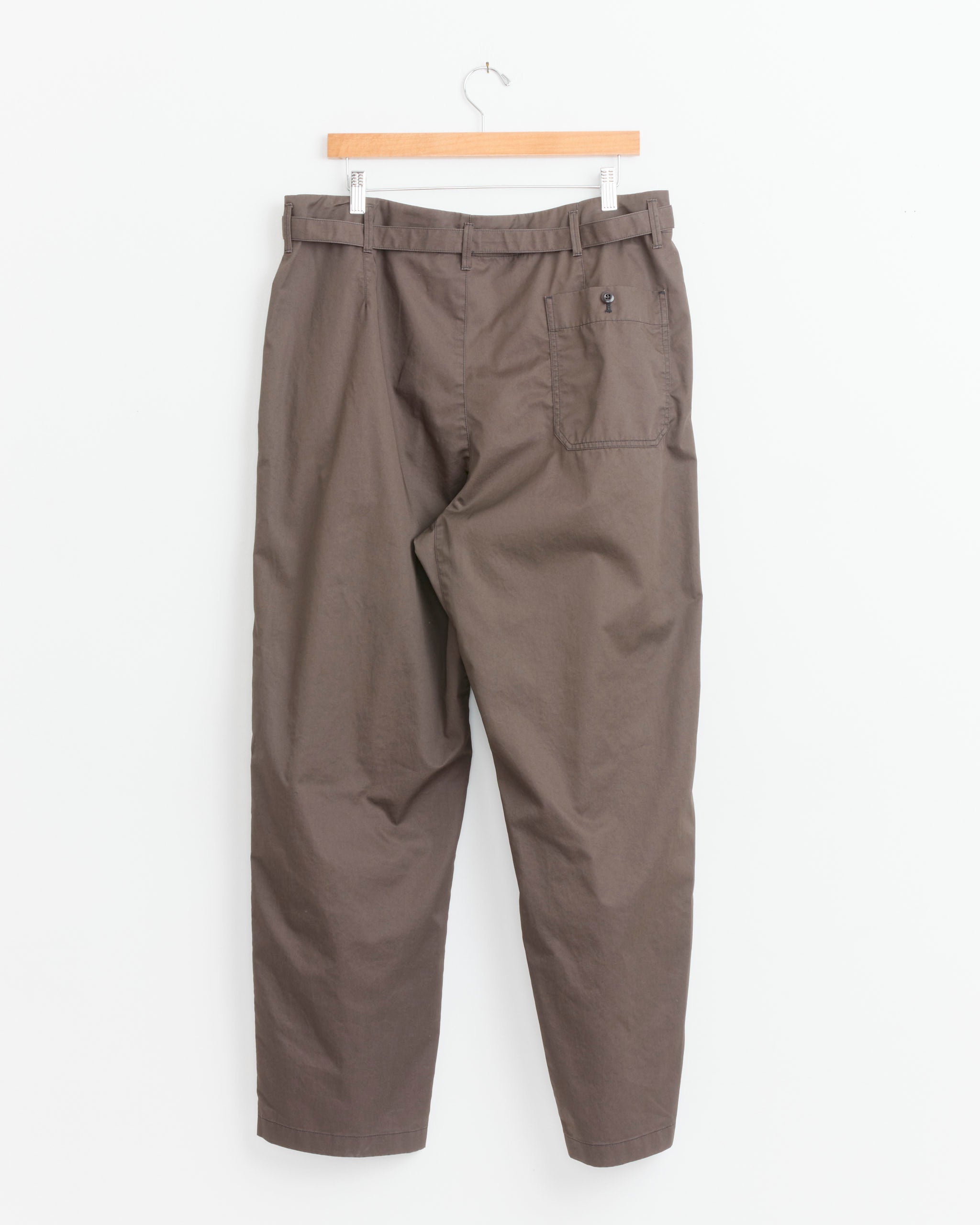The Lemaire Tapered Workwear Pant in Ristretto, crafted from brown cotton twill, hangs on a wooden hanger against a white background and features a right back pocket with button closure.