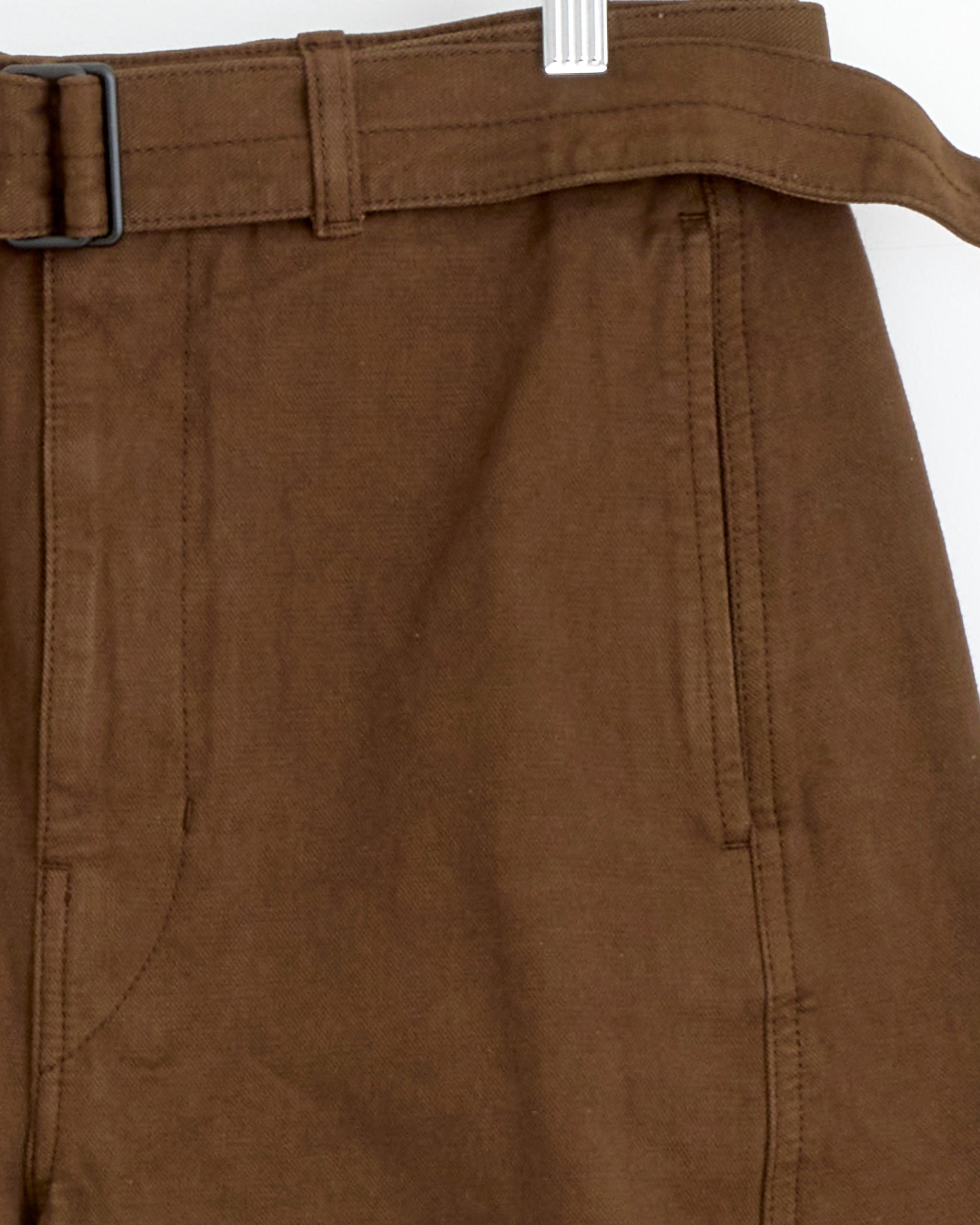 Close-up of the Lemaire Twisted Belted Pant in Cacao, featuring a belt, belt loops, and side pockets, hanging on a white hanger against a plain background. Inspired by Lemaire's minimalist style.