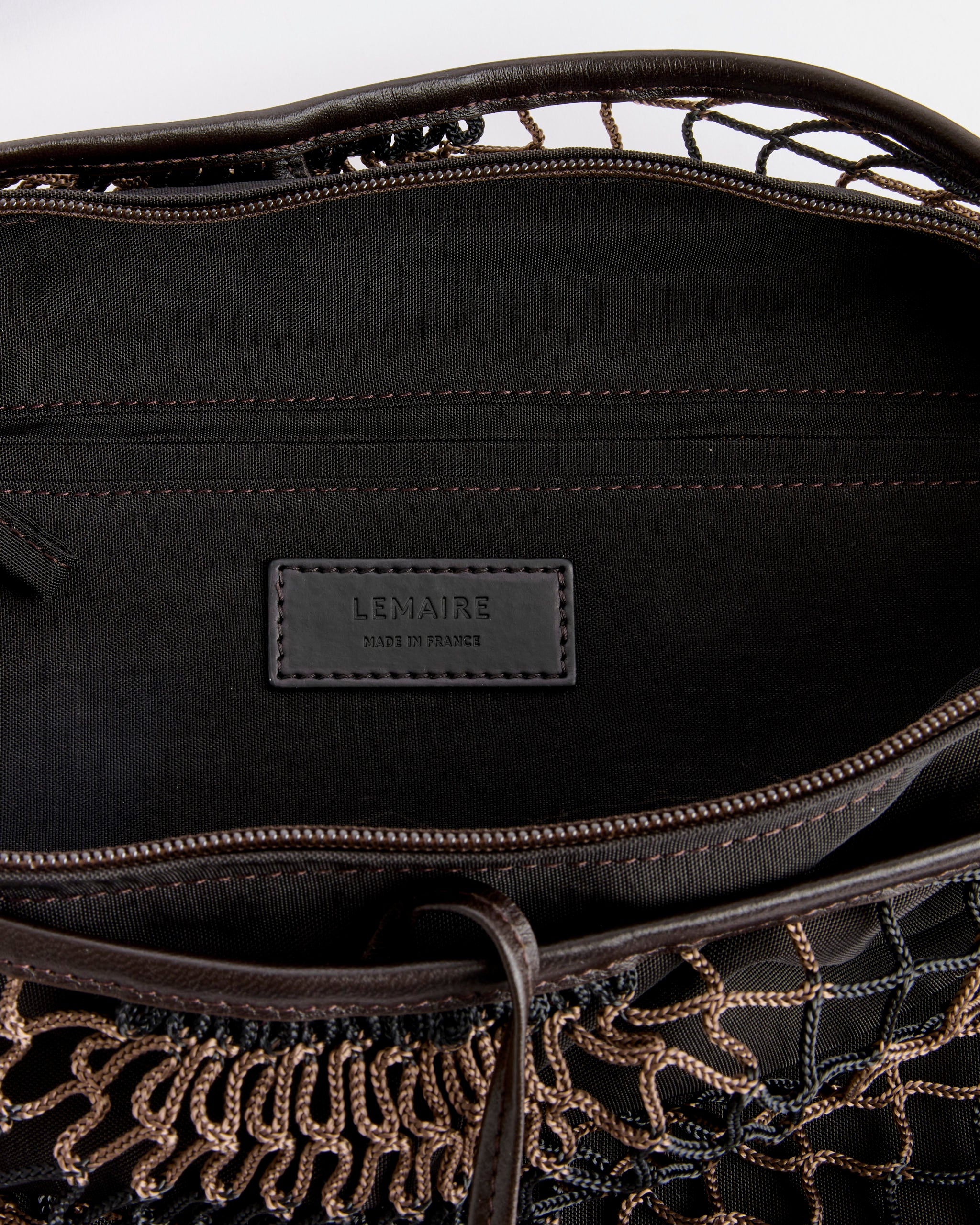 Close-up of the inside of a Lemaire Croissant Filt Bag in Chevron Marron Glace/Black, showing a stitched leather LEMAIRE label and visible tan thread details along the zippered opening and woven exterior.