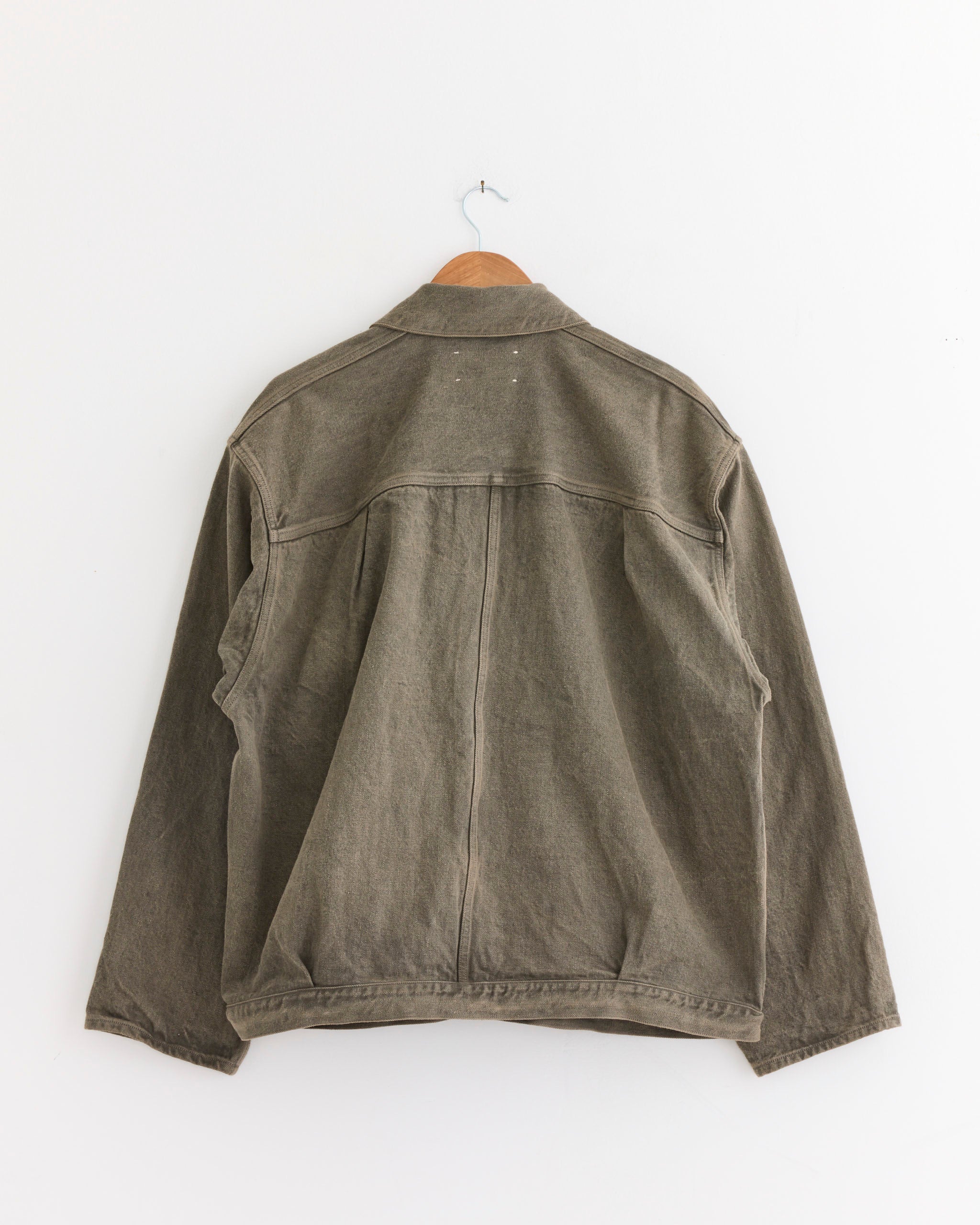 The Yoko Sakamoto Pleats Jacket in Mugwort, dyed with natural mugwort and made in Japan, hangs on a wooden hanger against a plain white wall, shown from the back.