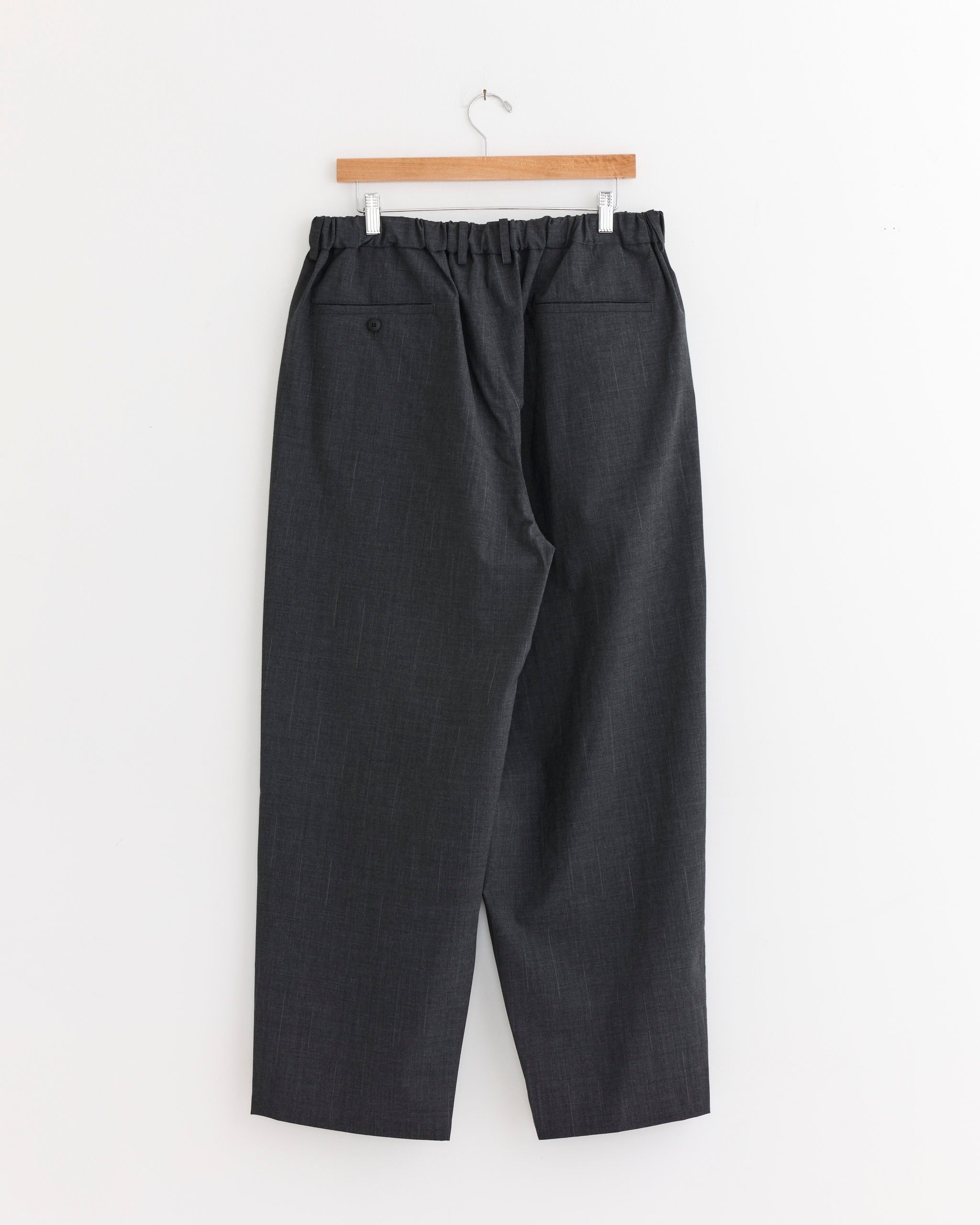 The Double Pleated Wide Easy Pant in Charcoal Grey by Yoko Sakamoto, made in Japan from 100% wool, features an elastic waistband and two back pockets (one with a button), hanging on a wooden hanger against a white wall.
