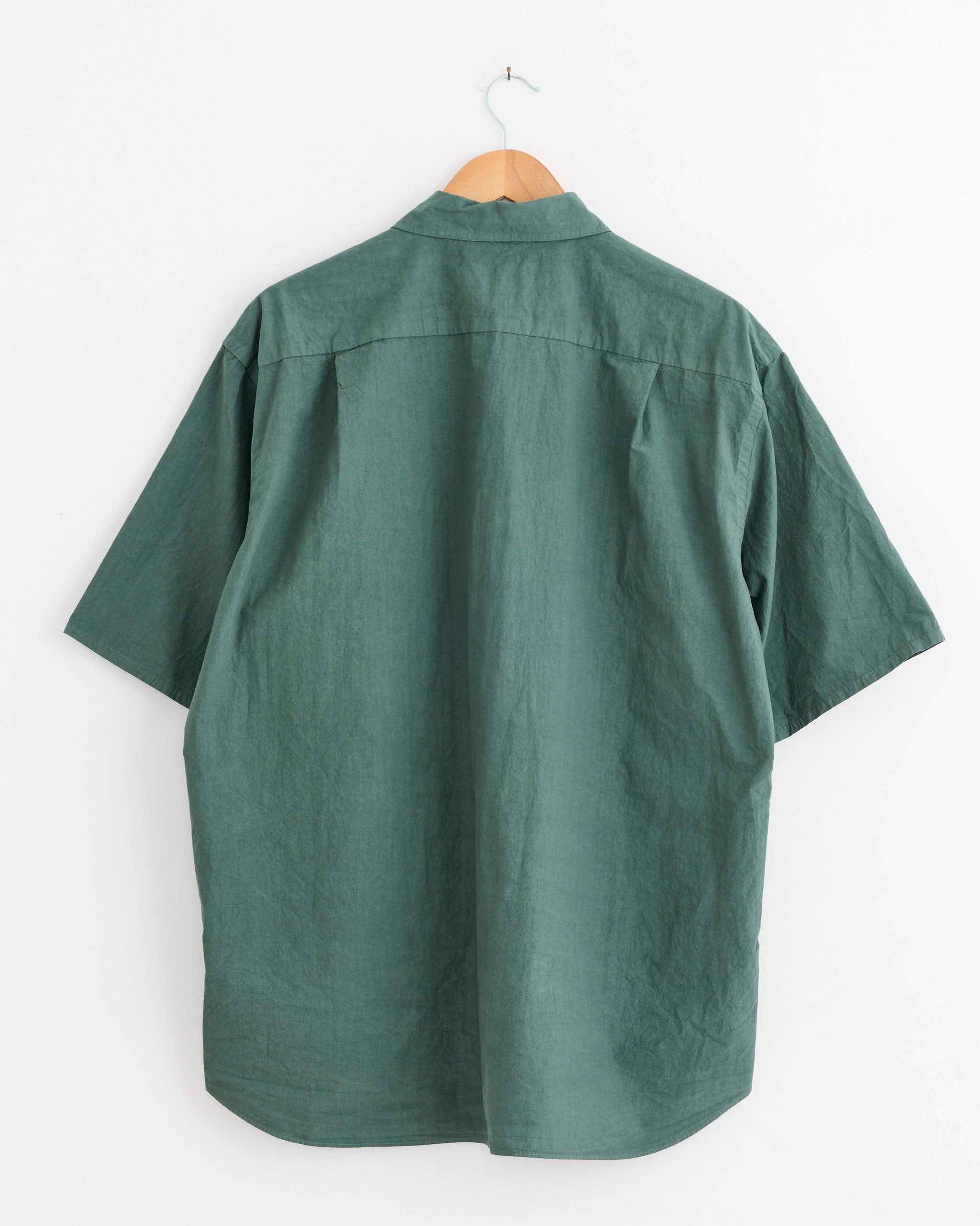 The Yoko Sakamoto Regular Collar S/S Shirt in Green, shown from the back, features short sleeves and a relaxed fit. It hangs on a wooden hanger against a plain white wall.