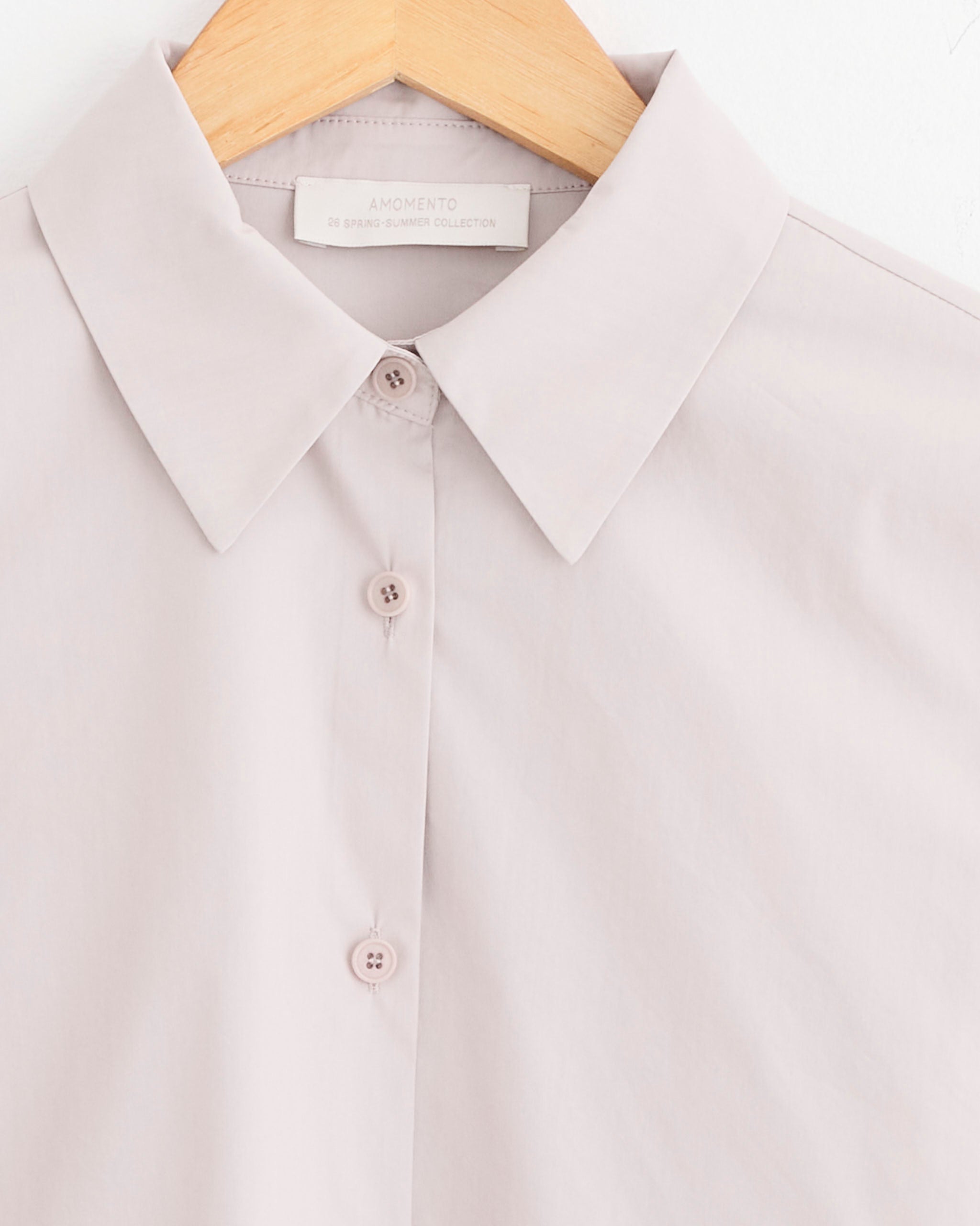 A close-up of the Slim Shirt in Beige by Amomento shows its cotton-polyester fabric, collar, upper chest, and three buttons on a wooden hanger. For the perfect fit, refer to our size guide.