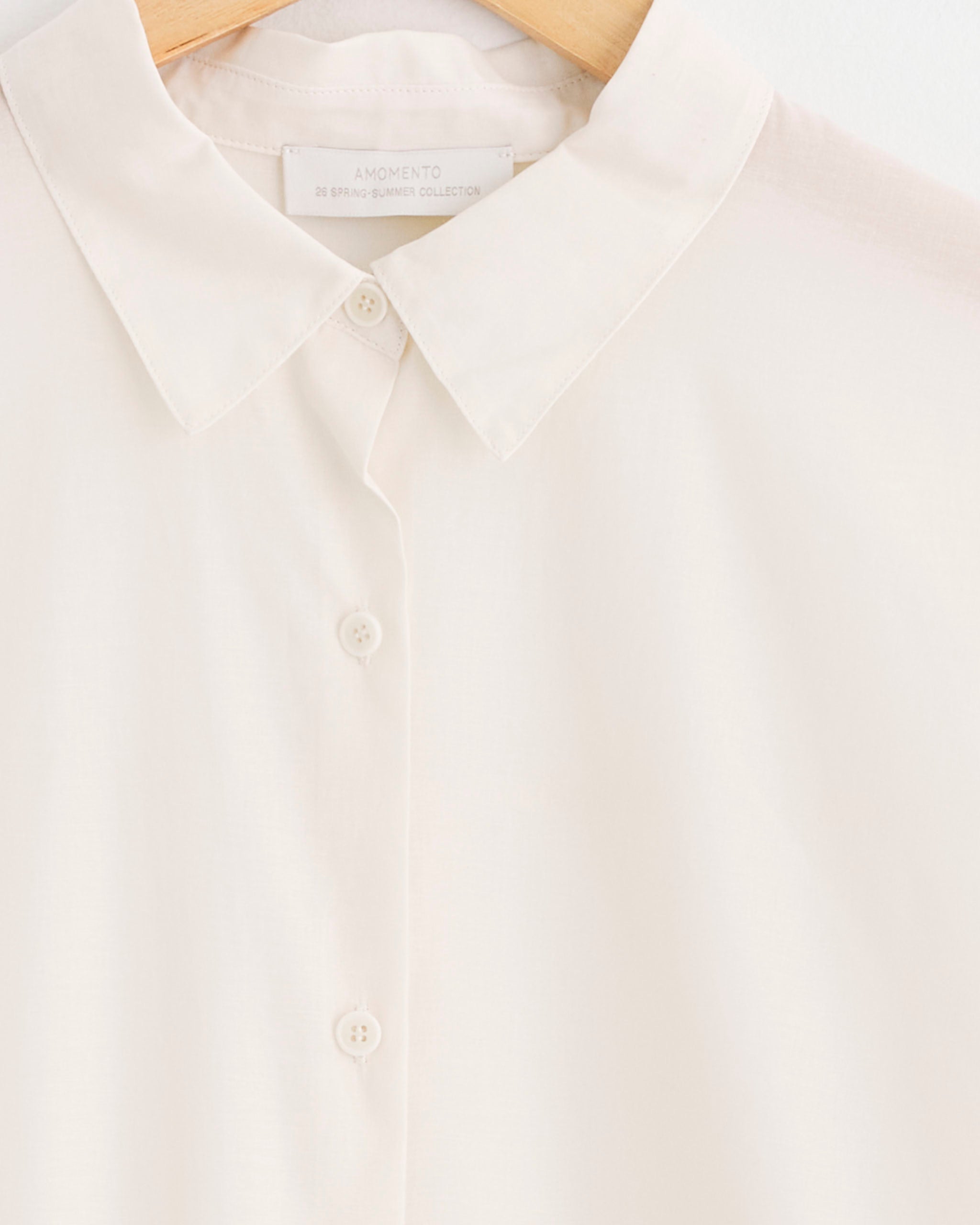 A close-up of the Sheer Cotton Classic Shirt in Ivory by Amomento, made from light cotton, on a wooden hanger, highlighting the collar, three buttons, and part of the inner brand label.