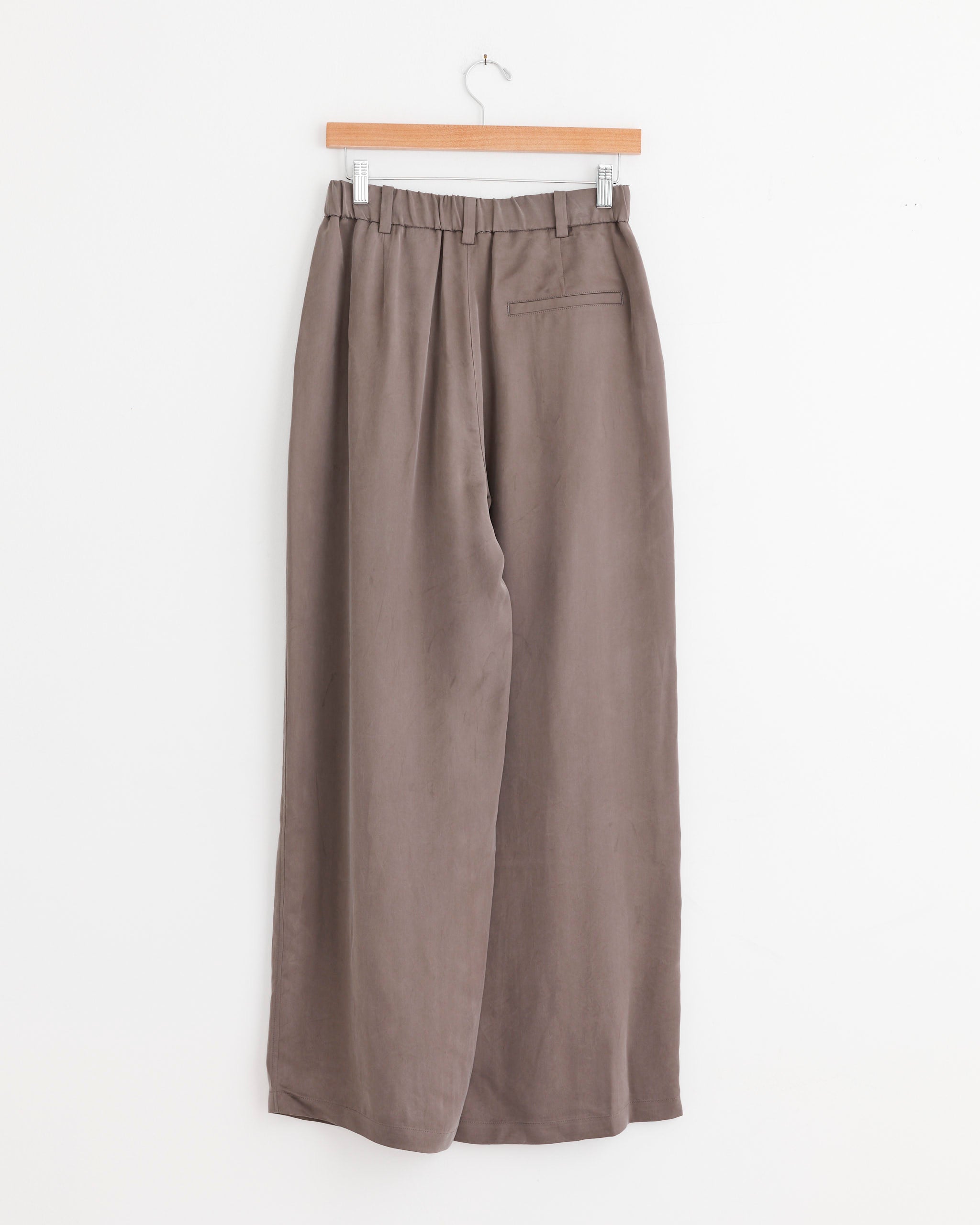 Amomento's Two Tuck Wide Pant in Grey, featuring wide legs, pleats, an elastic waistband, and a back pocket, hangs on a wooden hanger against a plain white background.