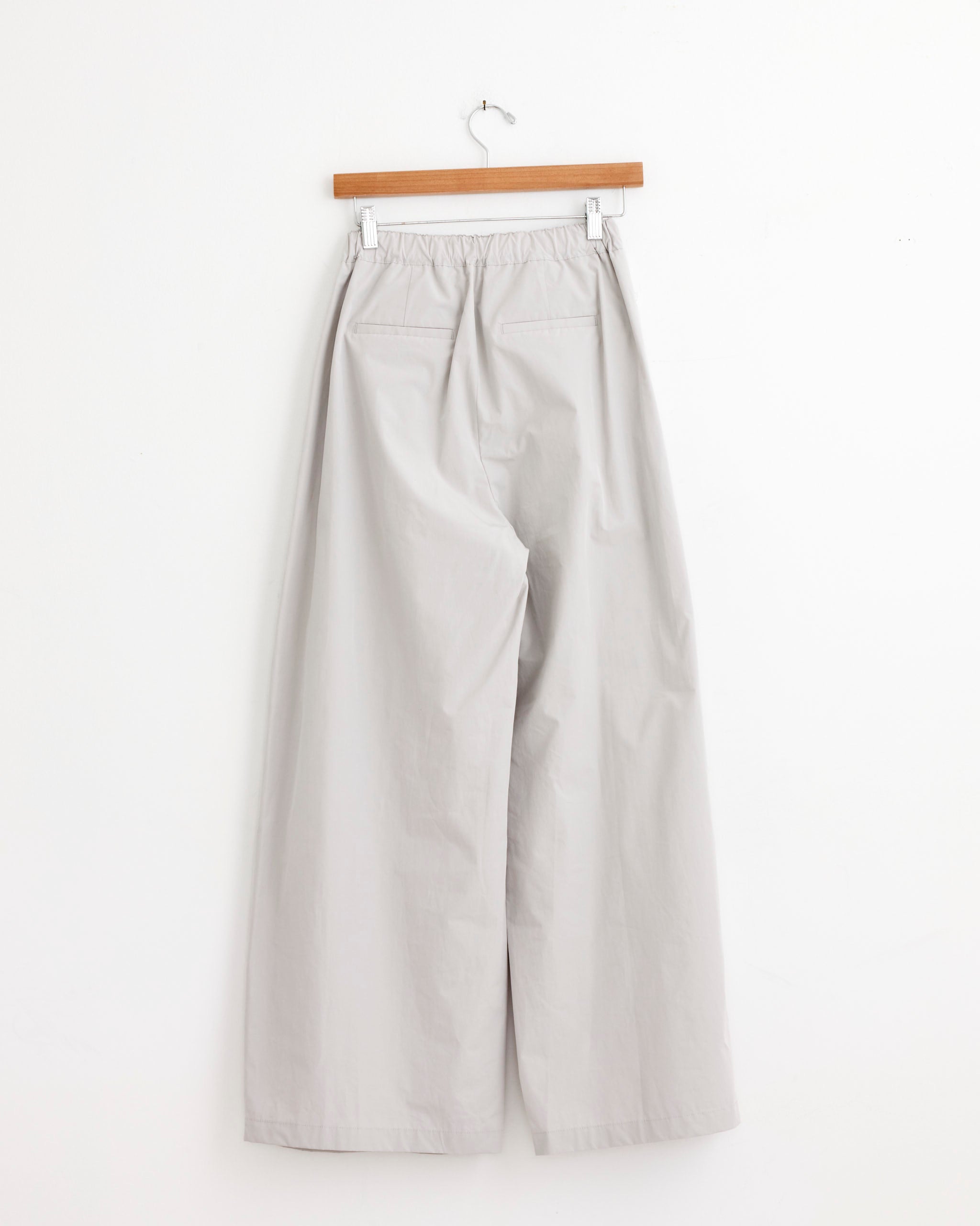 The Amomento Three Tuck Banding Pant in Grey offers a relaxed, wide fit with an elastic waistband, lightweight fabric, and two back pockets. Shown hanging on a wooden hanger against a plain white wall.