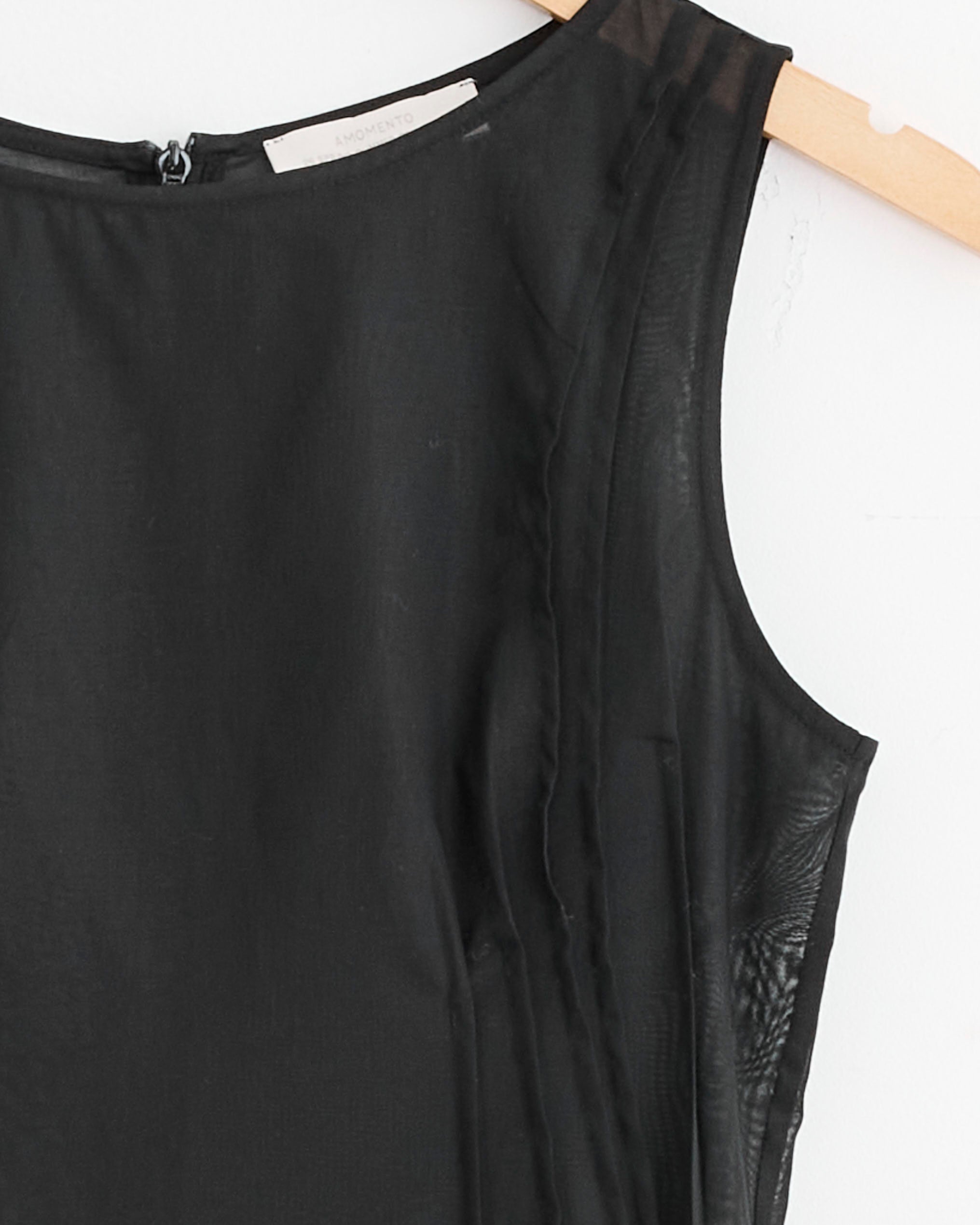 The Amomento Pintuck Dress in Black is displayed on a wooden hanger against a white wall, featuring sheer fabric details and a back zipper for a flattering fit. View sizing to find your perfect look.