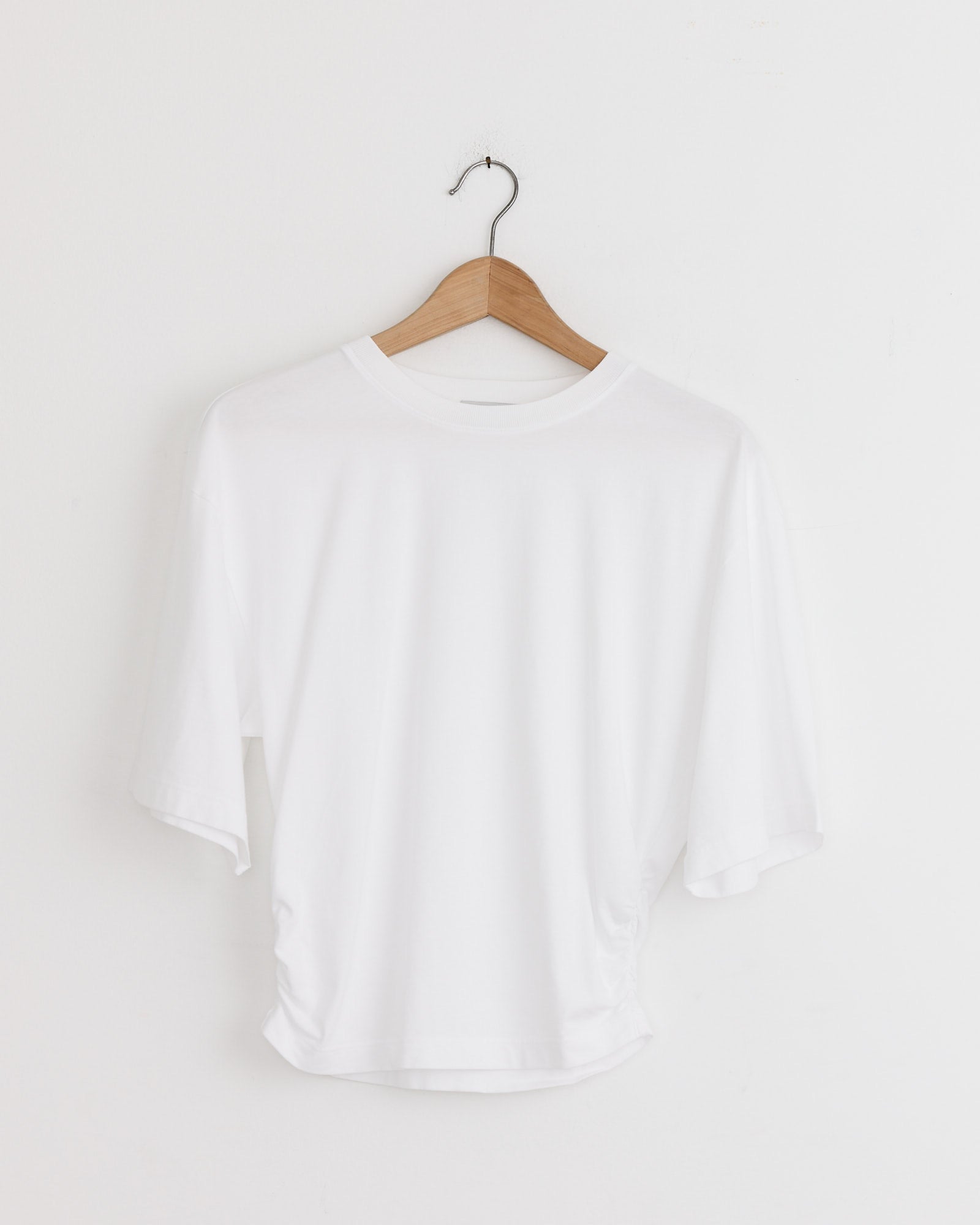 The Shari Tee in White by AGOLDE hangs on a wooden hanger against a white wall—perfect for any fit. Need sizing help? Chat with us online.