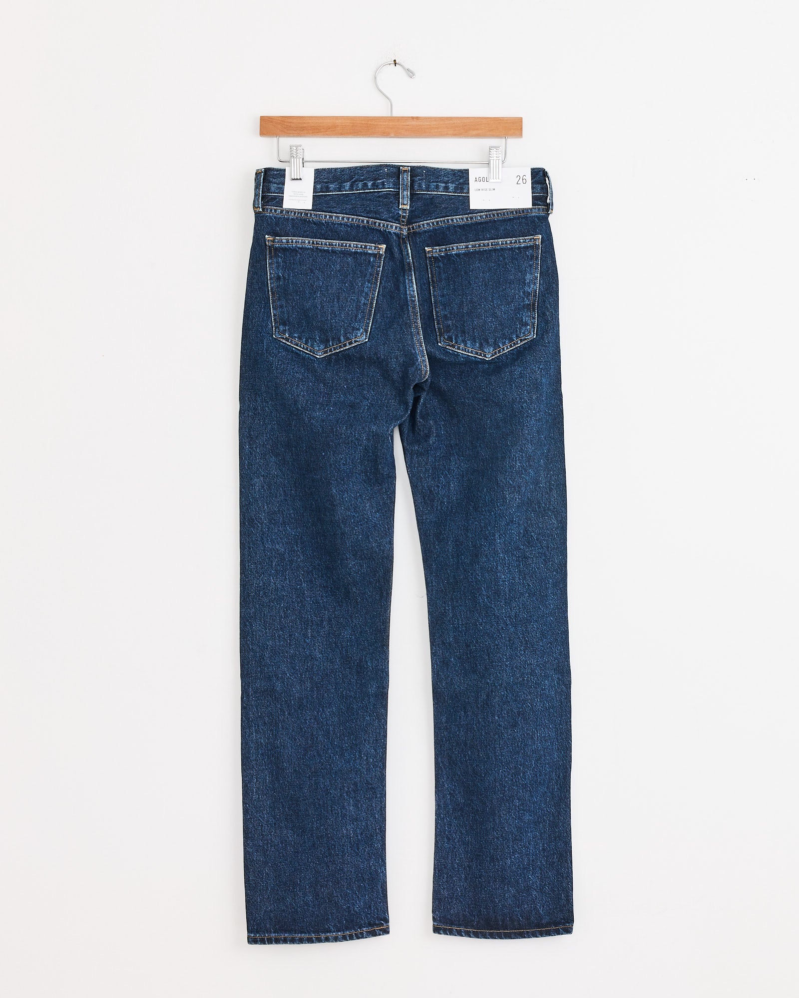 The AGOLDE Low Rise Slim in Pendulum jeans hang on a wooden hanger against a white background, shown from the back to highlight the two rear pockets and straight-leg cut—ideal for those wanting classic style and accurate sizing.