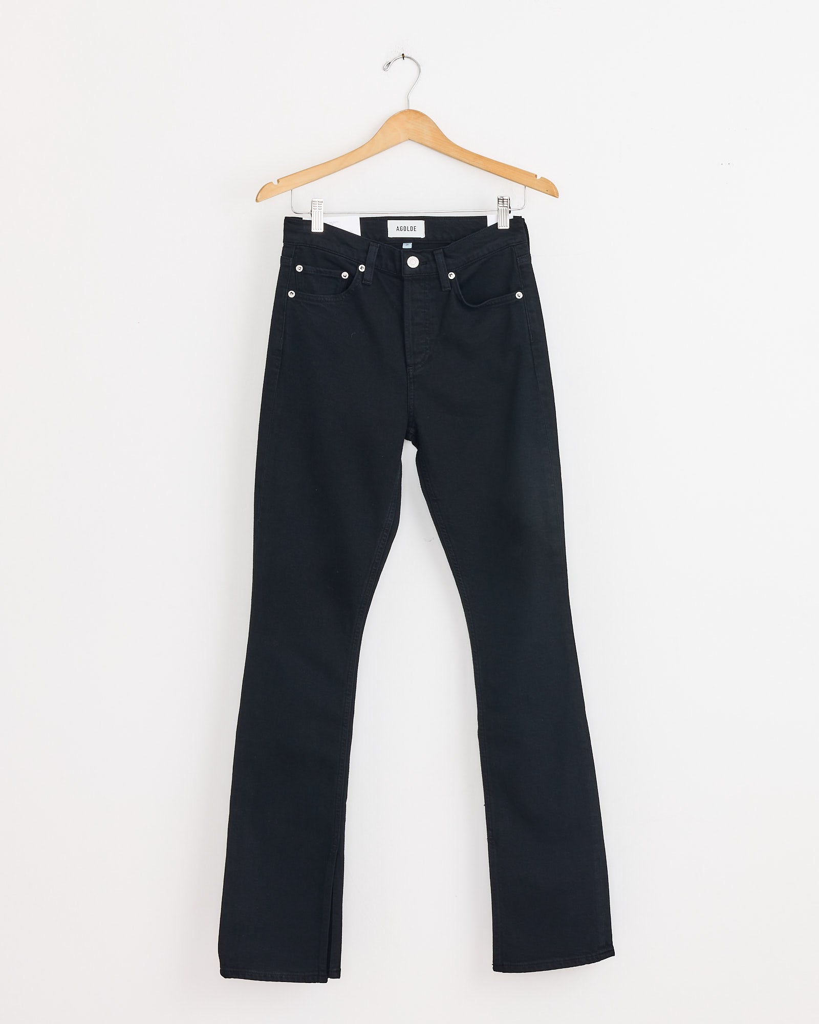 The AGOLDE Ingram Split Skinny in Immersion, featuring a classic five-pocket design and skinny legs, hangs on a wooden hanger against a white wall. Use our online chat for sizing questions.