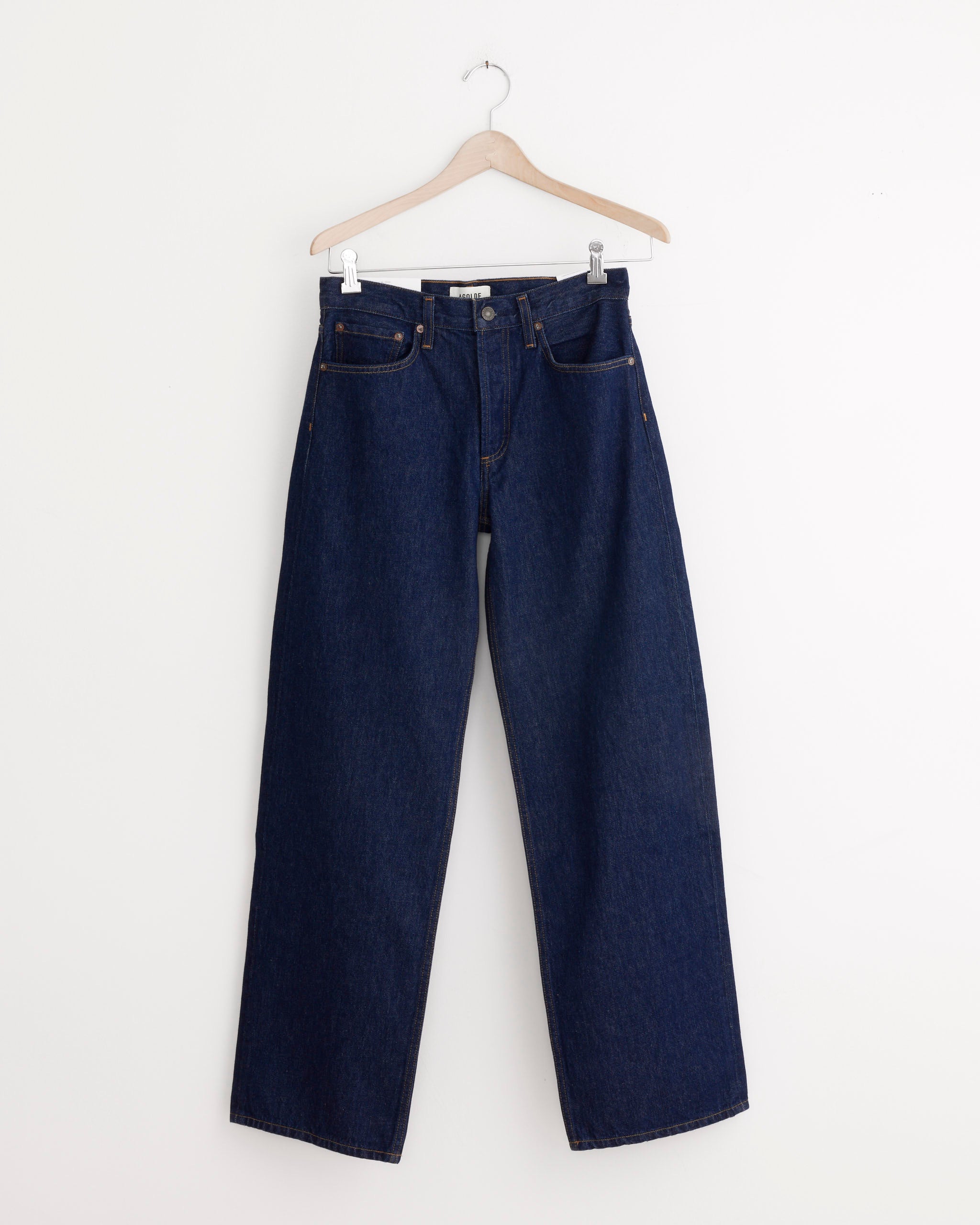 The AGOLDE Arc Jean 32" in Rinse, a pair of dark blue straight-leg jeans made from regenerative cotton, hangs on a wooden hanger against a plain white wall.