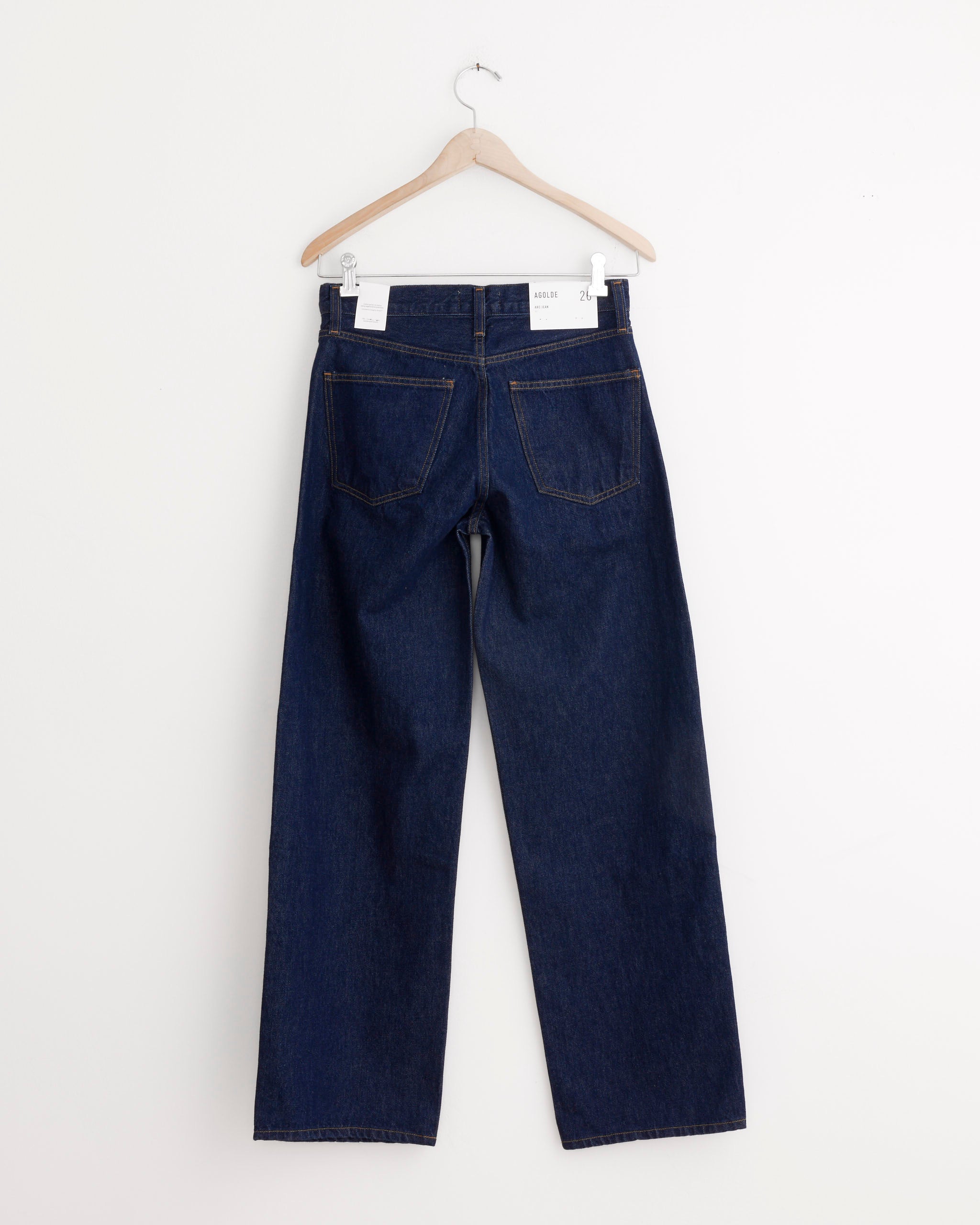 The AGOLDE Arc Jean 32" in Rinse, featuring wide legs, two back pockets, and a white waistband label, hangs on a wooden hanger against a plain white wall.