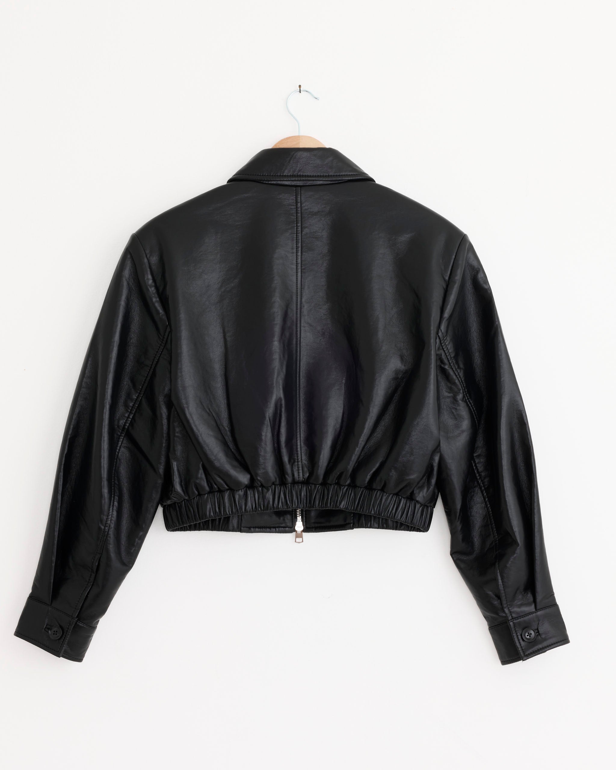 The AGOLDE Nahla Cropped Jacket in Detox features a collar, long sleeves, elastic waistband, and buttoned cuffs. The black leather jacket is shown from the back as it hangs on a wooden hanger against a plain white wall.