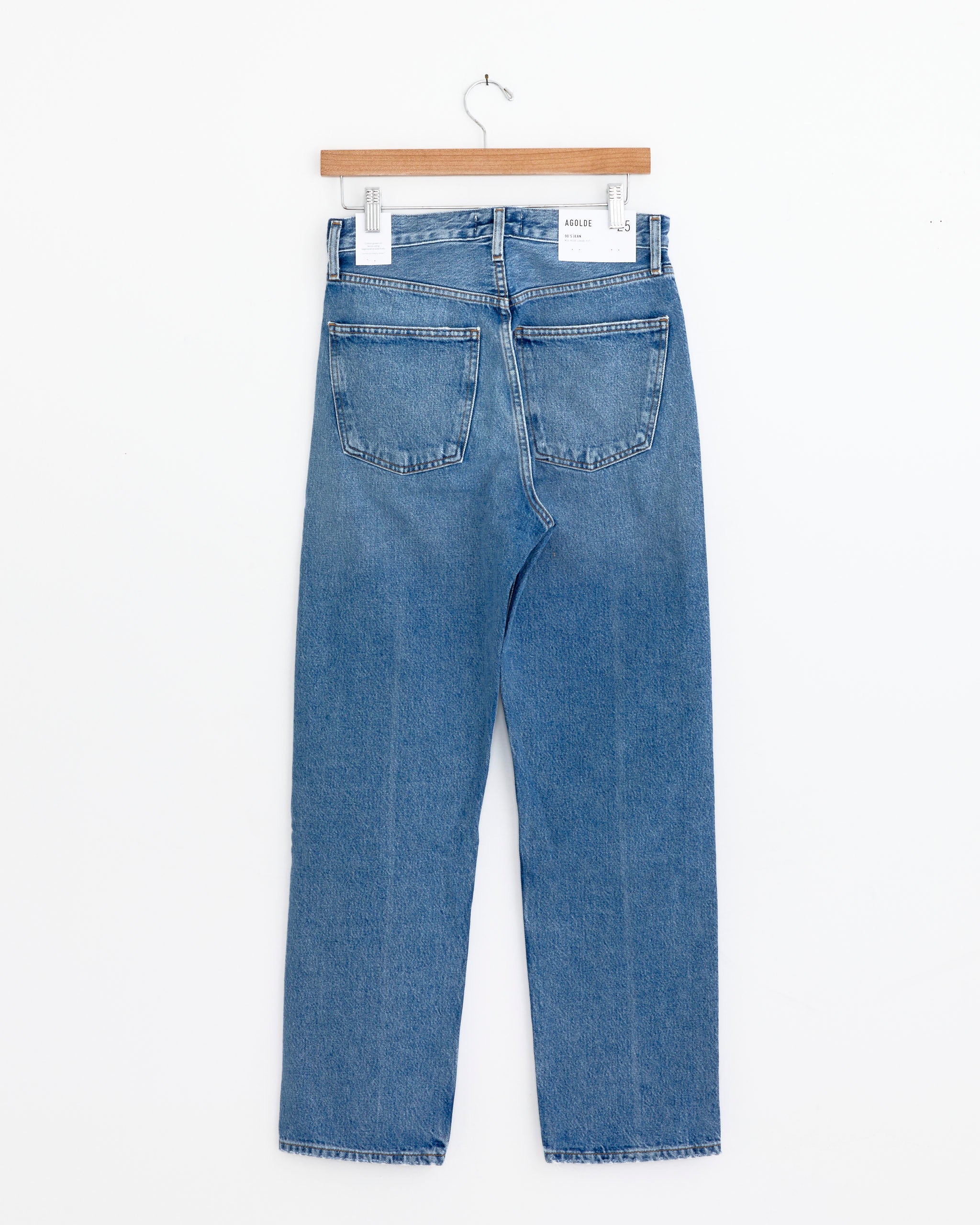 A pair of AGOLDE 90's Jean in Vault, made from regenerative cotton denim, hangs on a wooden hanger against a white background, shown from the back with two rear pockets and a white waistband label.