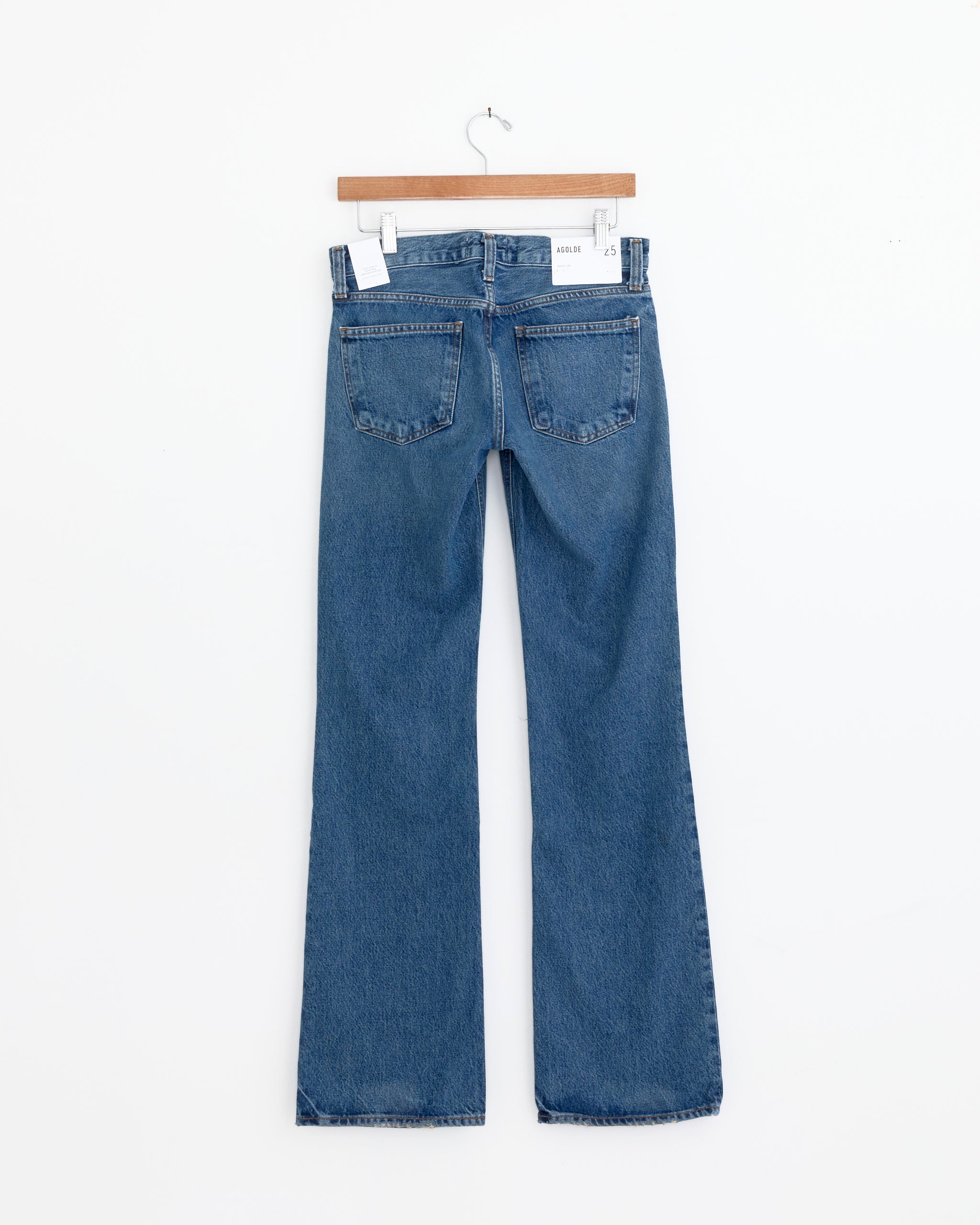 The AGOLDE Low Rise Boot in Margin, a pair of indigo wash bootcut jeans, hangs on a wooden hanger against a plain white wall, displayed from the back with two rear pockets visible.