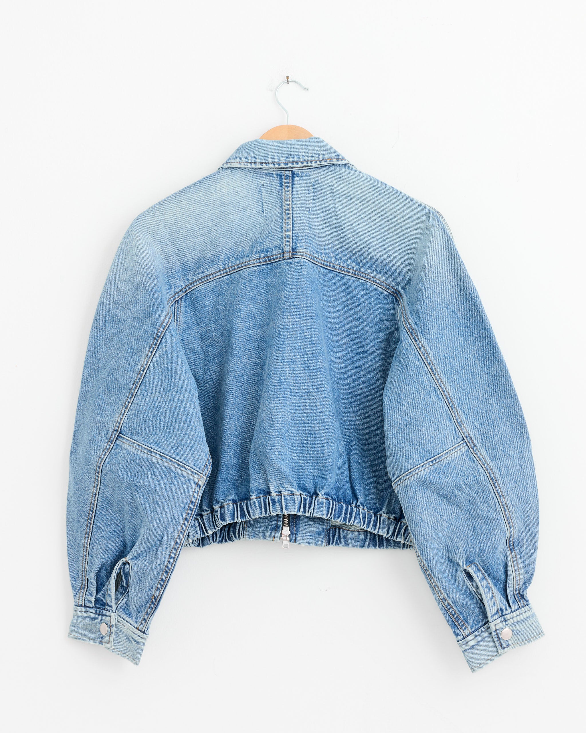 The Dixon Cocoon Bomber Jacket in Outbreak by AGOLDE, a light blue denim jacket with long sleeves, button cuffs, and an elastic waistband, hangs on a wooden hanger against a plain white background, shown from the back.