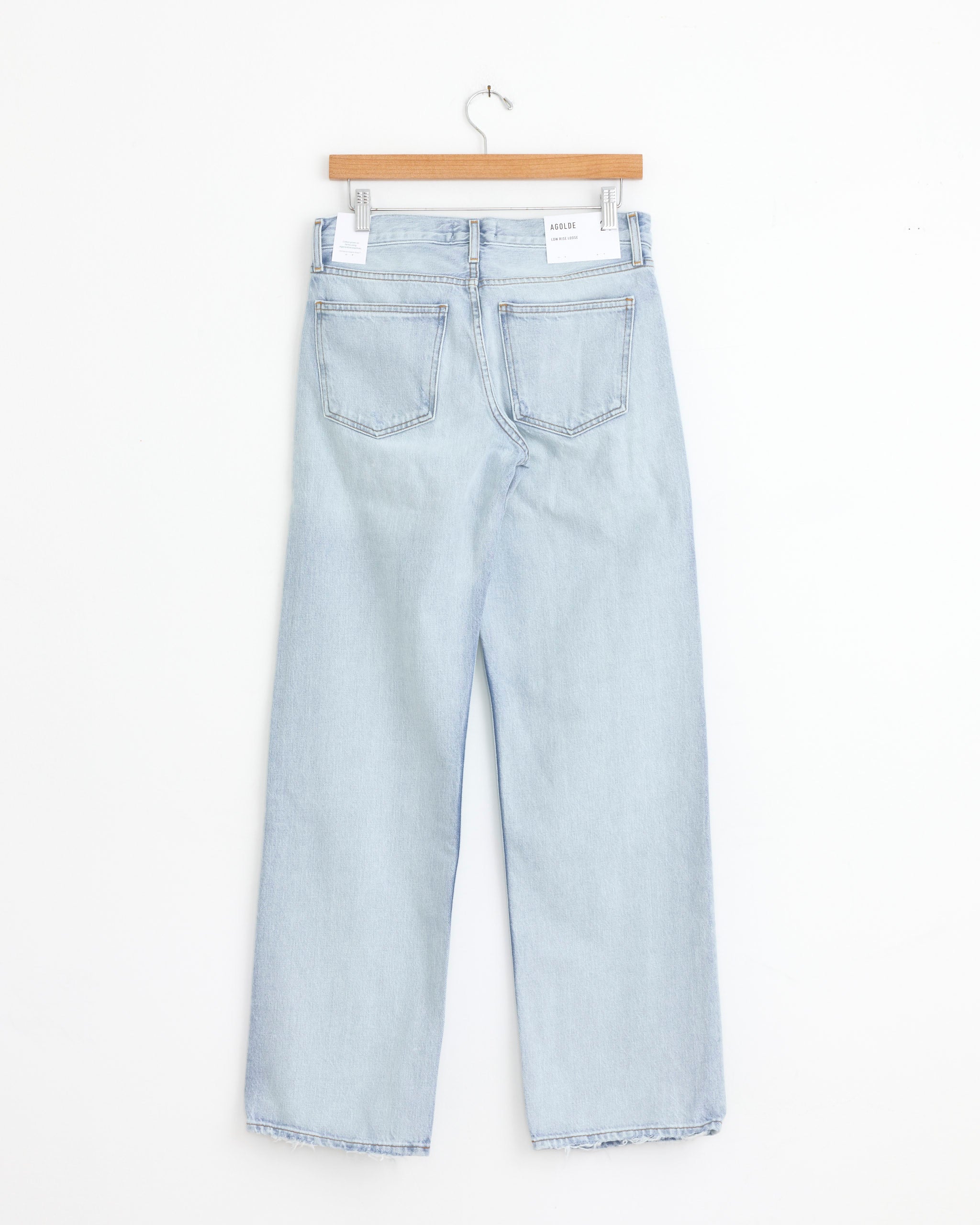 The AGOLDE Low Rise Loose in Amplify jeans, featuring a light blue wash and straight leg cut, are displayed hanging on a wooden hanger with the back and two pockets visible against a white background.