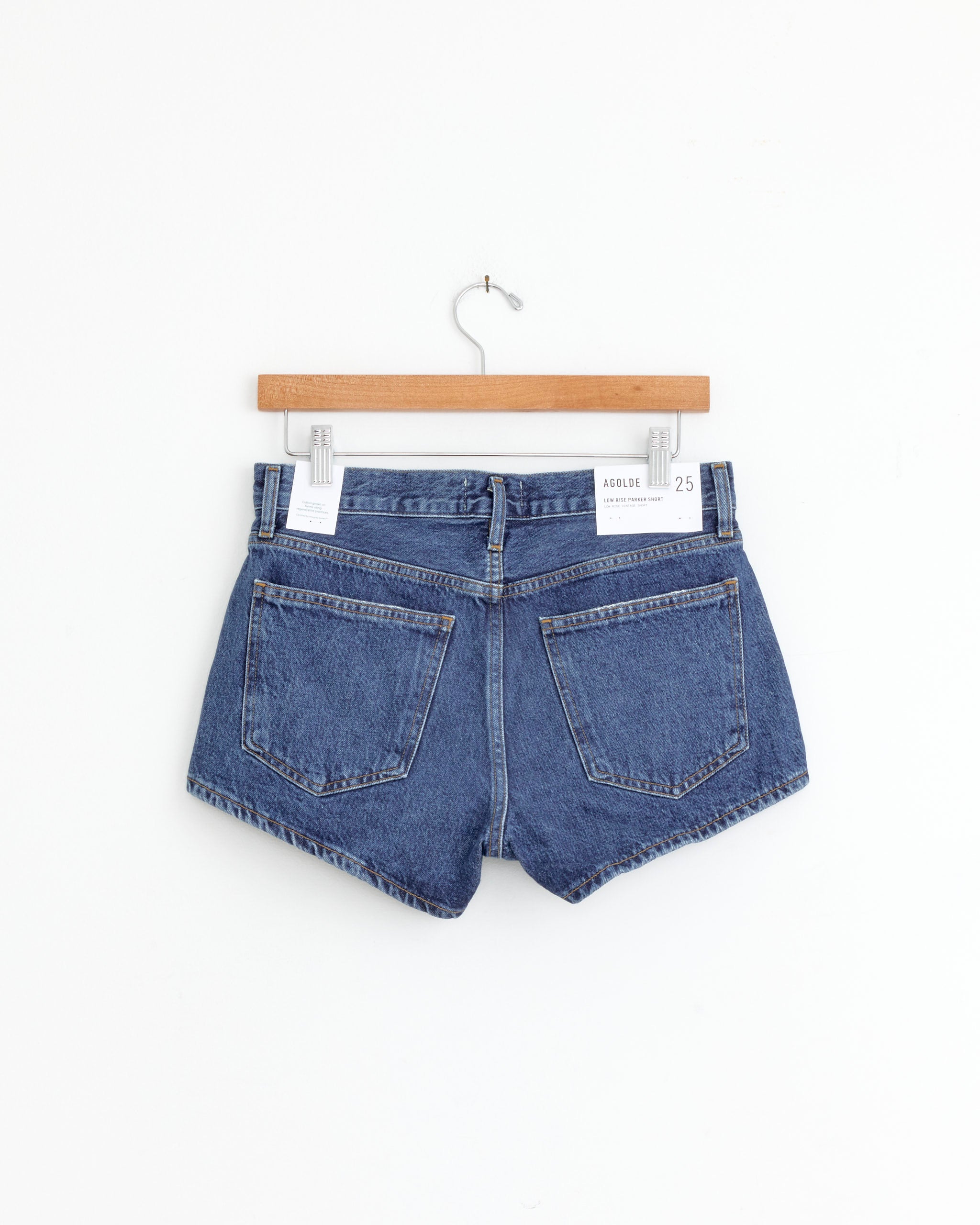 The AGOLDE Low Rise Parker Short in Enamour, a pair of blue denim shorts with large back pockets, is shown on a wooden hanger against a plain white background.