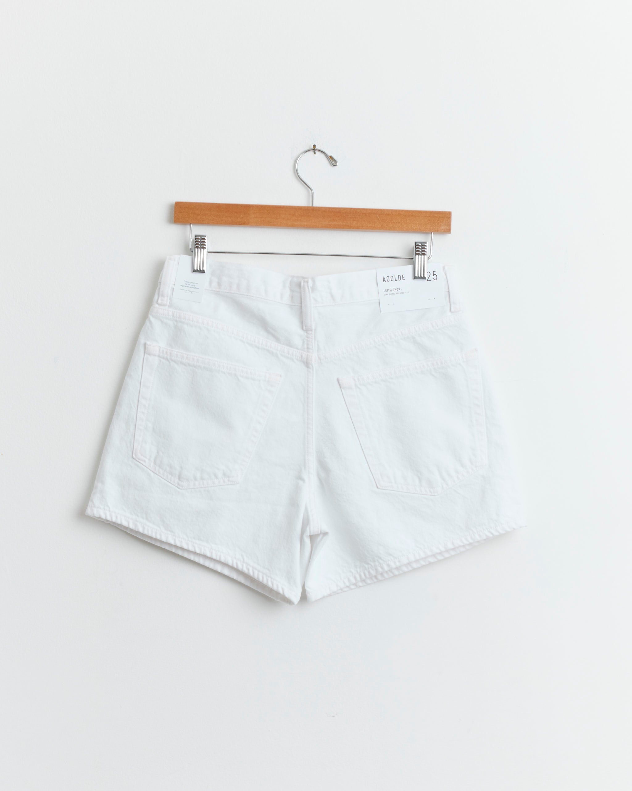 The AGOLDE Leith Short in Marshmallow, crafted from regenerative cotton, hangs on a wooden hanger against a white wall, showcasing its two rear pockets and label near the waistband.