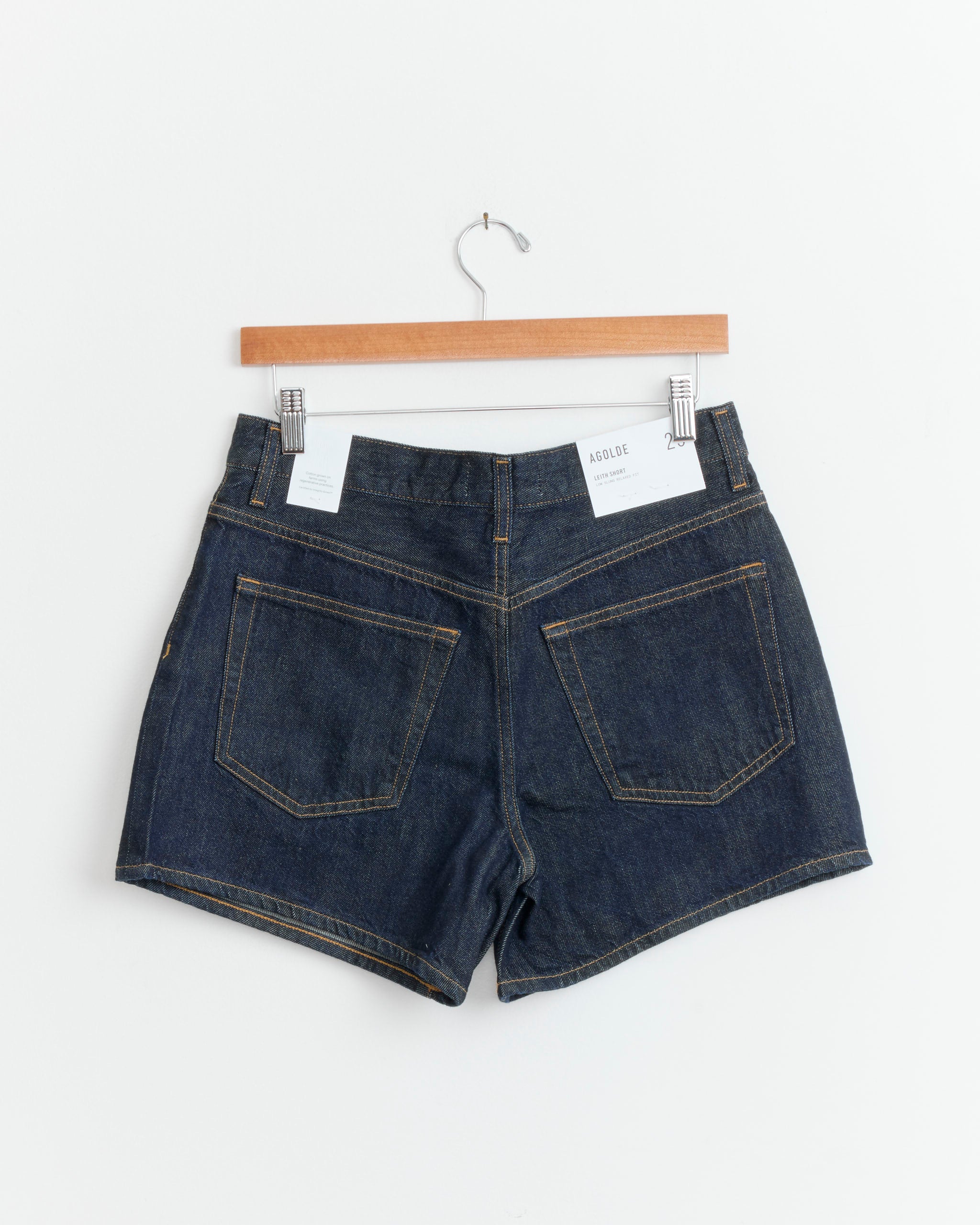 The AGOLDE Leith Short in Rinse is an oversized denim short made from regenerative cotton with brown stitching and two back pockets, shown hanging on a wooden hanger against a plain white background.