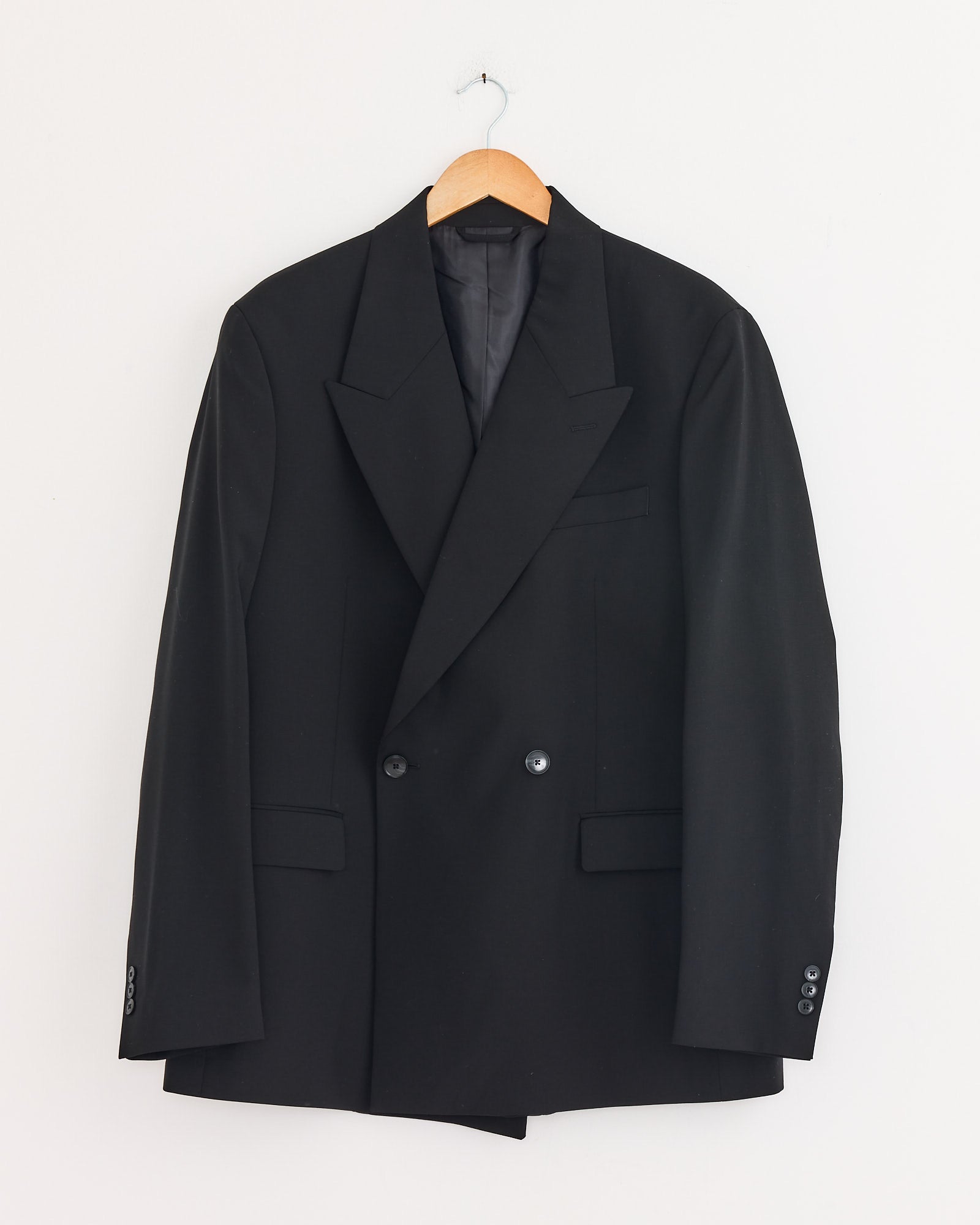 The mfpen Double Breasted Blazer in Black hangs on a wooden hanger against a white wall. Made from Italian wool, it features peak lapels, two buttons, and front flap pockets.