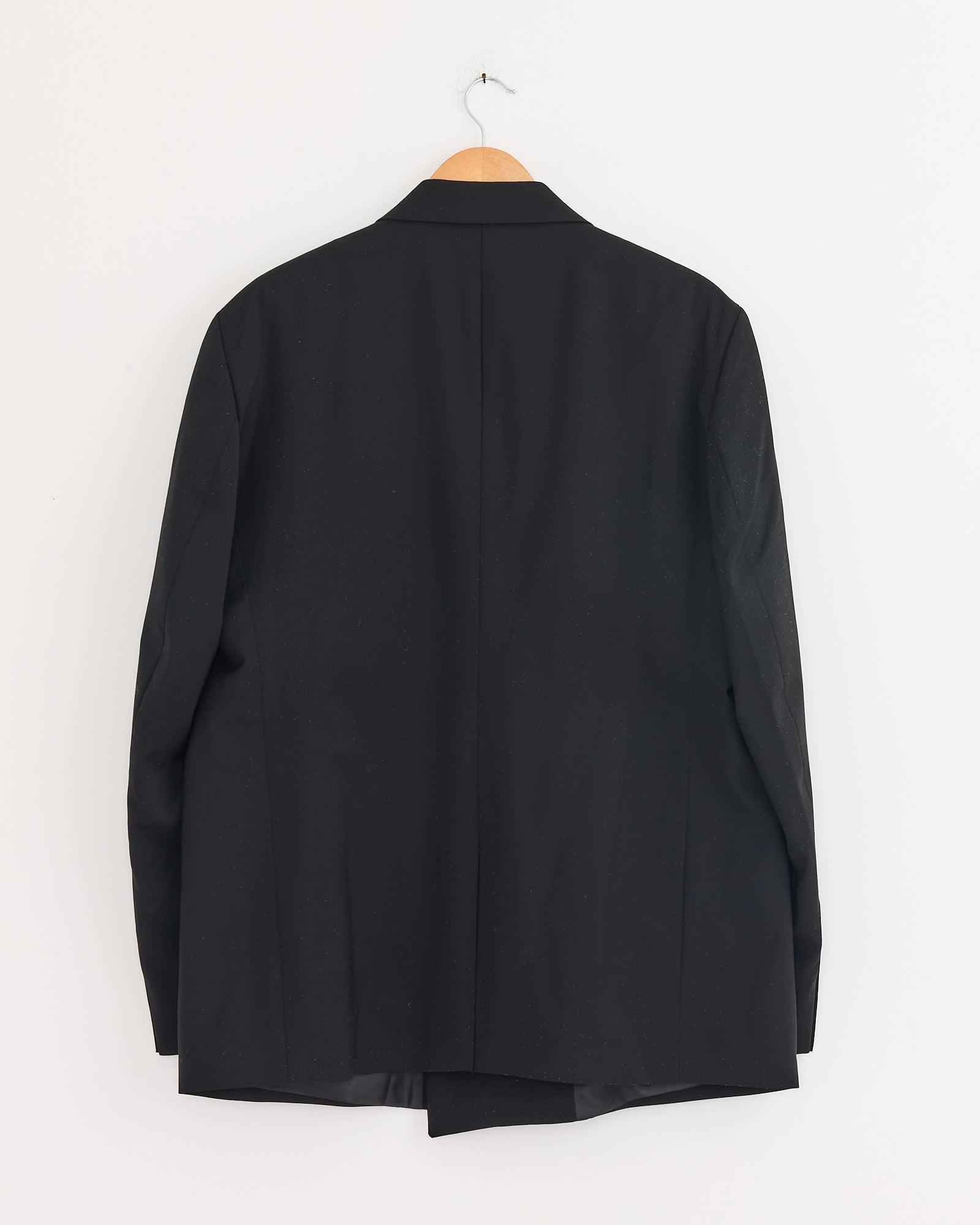 The mfpen Double Breasted Blazer in Black, crafted from Italian wool, is displayed on a wooden hanger against a plain white wall, shown from the back.