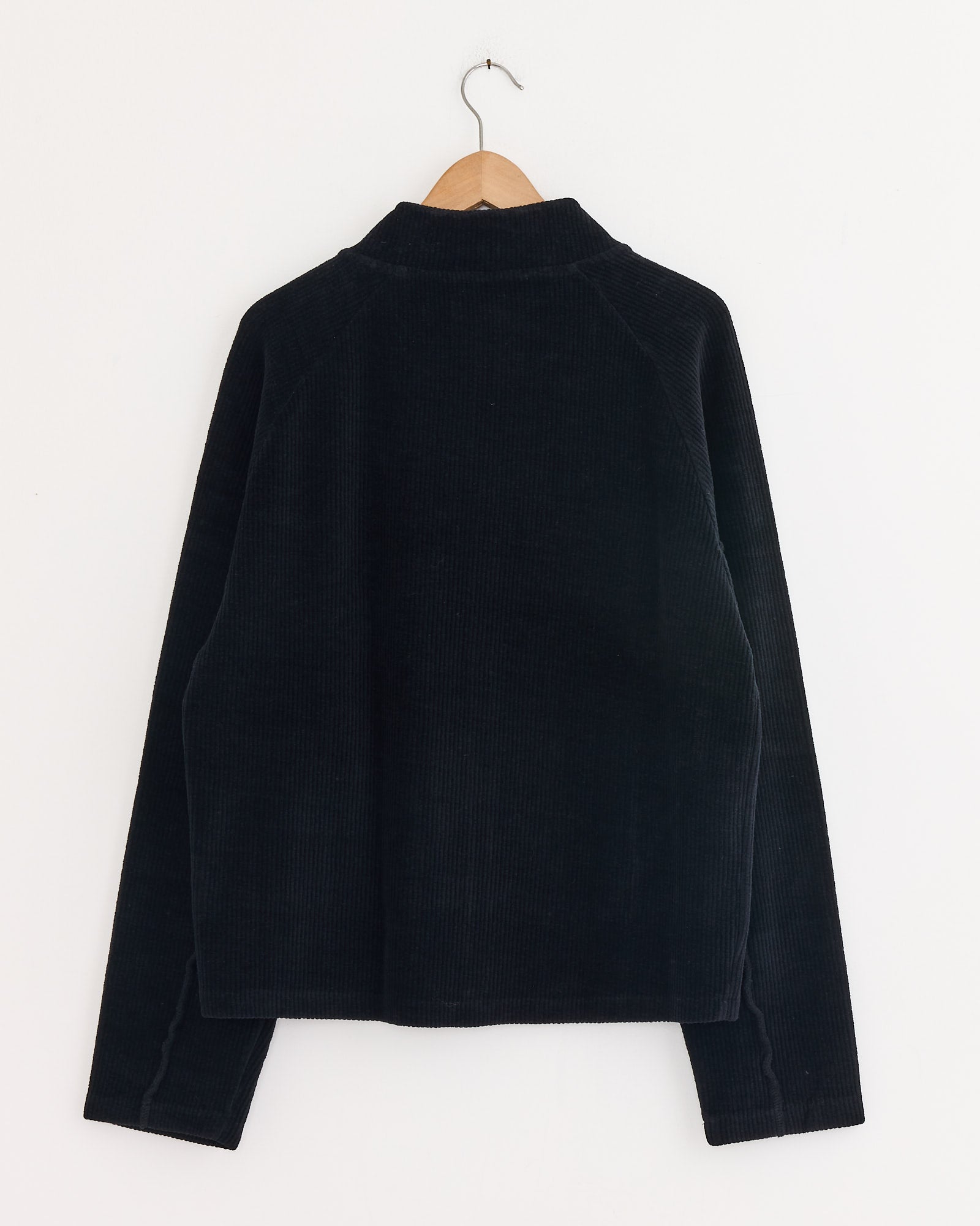 The mfpen Leisure Zip Up in Black, featuring a corduroy-like texture, long sleeves, and a collar, hangs on a wooden hanger against a plain white wall and is shown from the back.