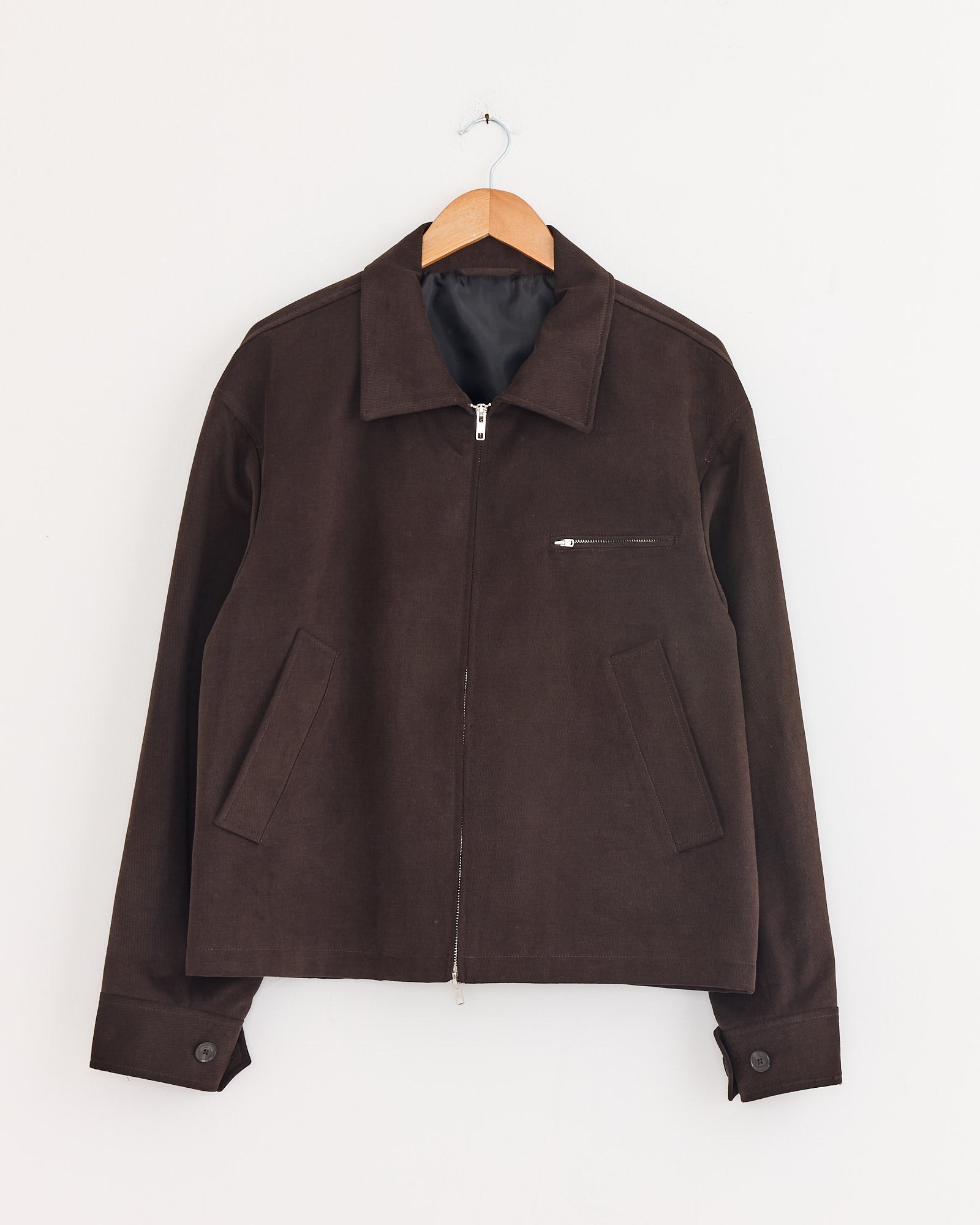 The mfpen Postal Jacket in Brown is a long-sleeve, Italian deadstock twill jacket with a collar, front zipper, two slanted side pockets, a zippered chest pocket, and buttoned cuffs—shown on a wooden hanger against a white wall.