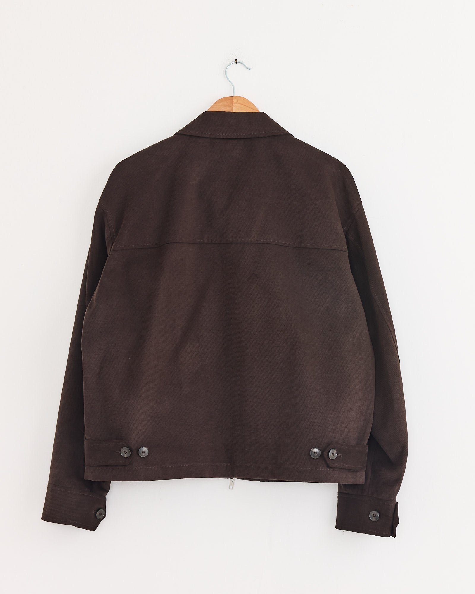 The mfpen Postal Jacket in Brown hangs from a wooden hanger against a plain white wall, shown from the back with button details on the lower hem and cuffs.