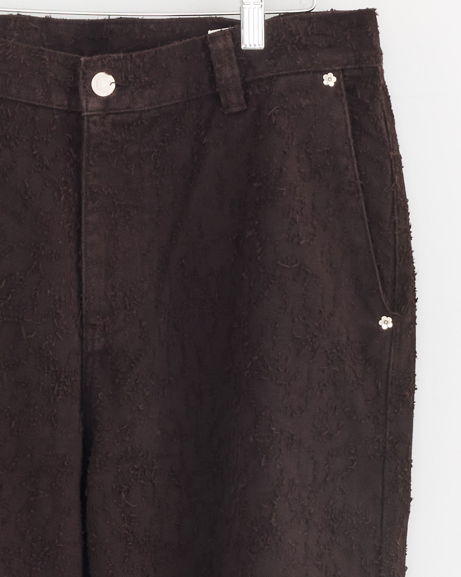 Close-up of the upper half of mfpen Punch Out Jean in Dark Brown, made from organic cotton denim, shown on a hanger with button closure, belt loops, and floral-shaped metal rivets on the pockets.